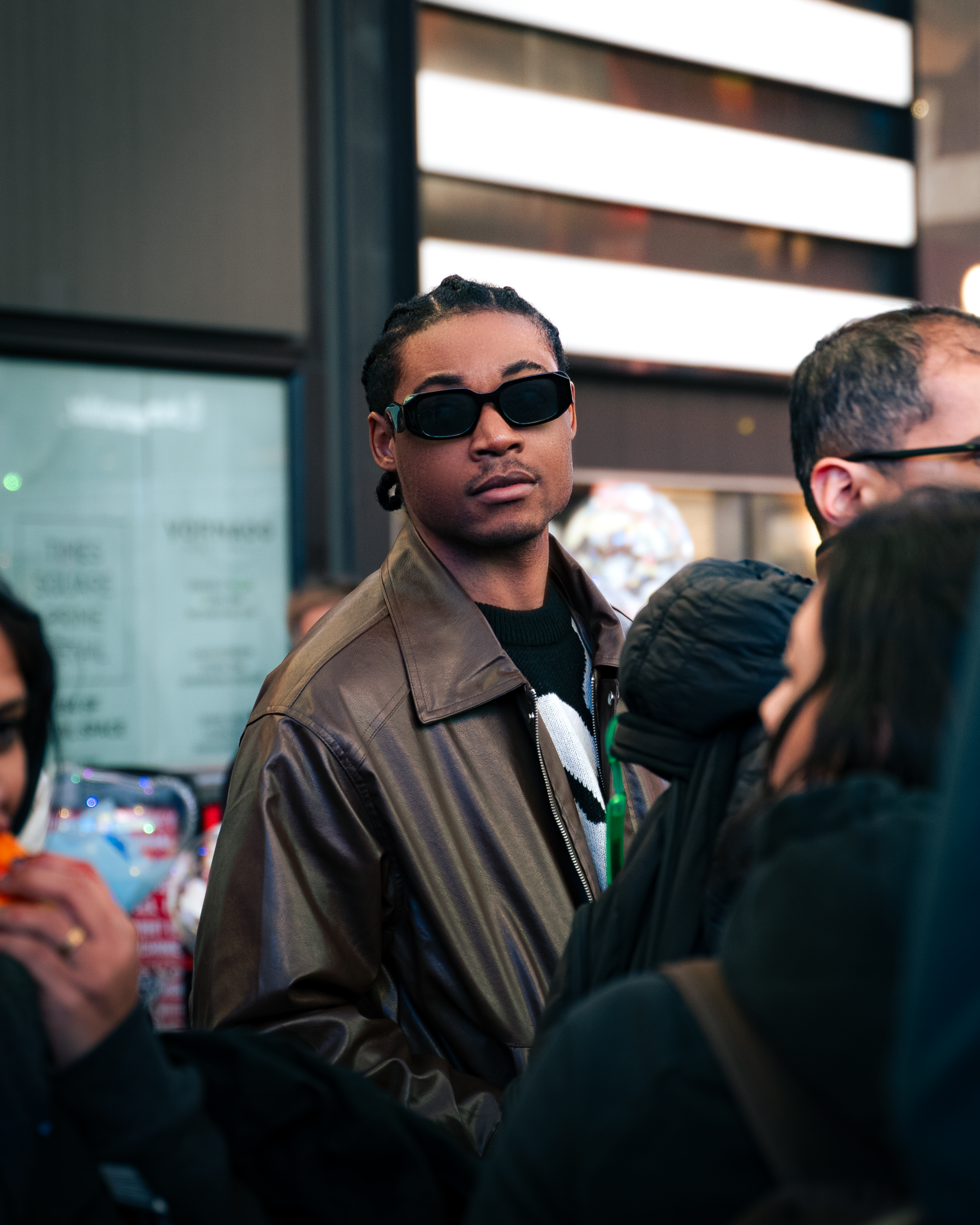 A young man with dark hair in bantu knots, wearing dark sunglasses, a brown leather jacket, and a black sweater with white stripes, standing among a crowd.