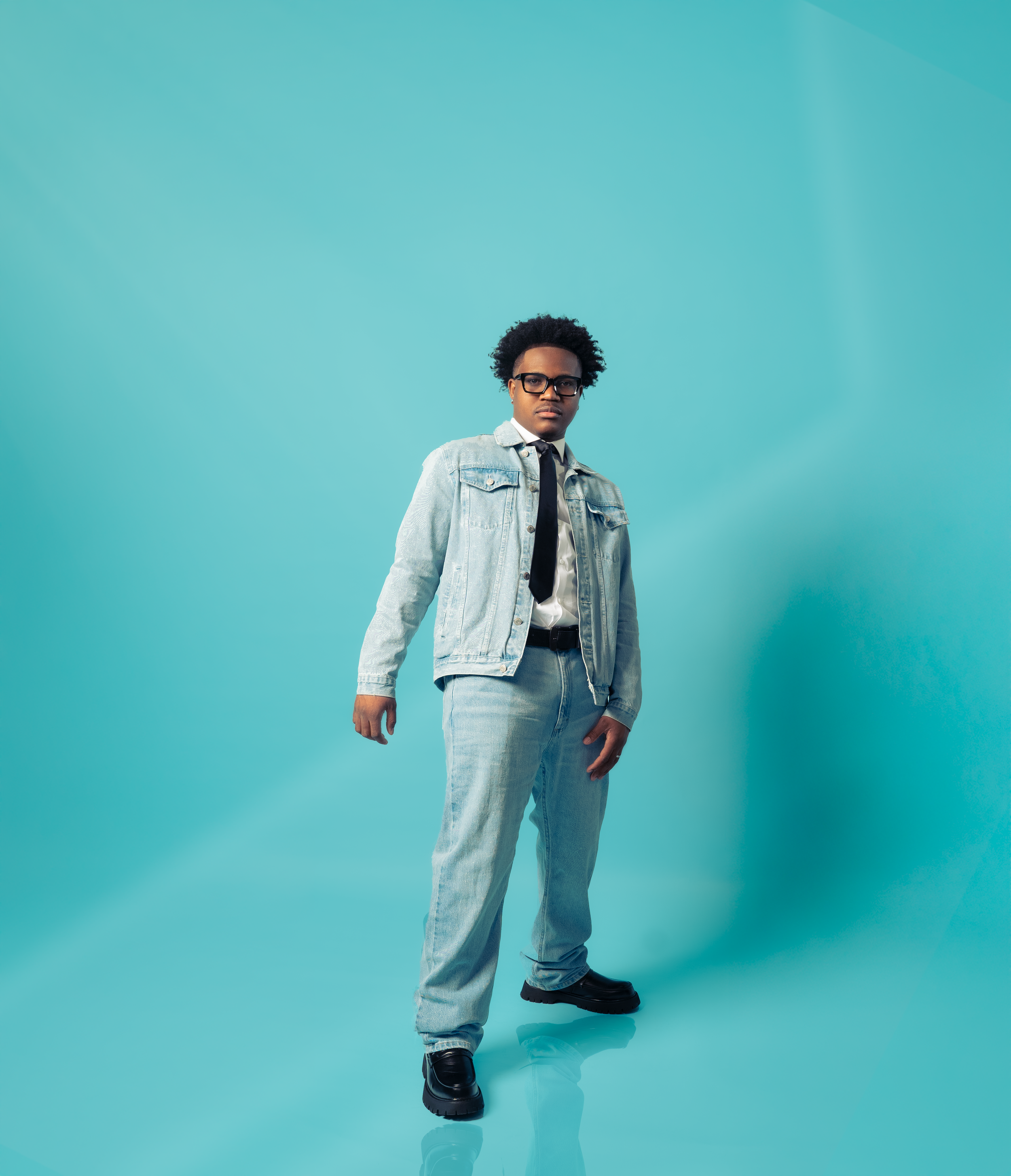 A young man with curly hair wearing glasses, a denim jacket, white shirt, black tie, light blue jeans, and black boots standing against a plain teal background.