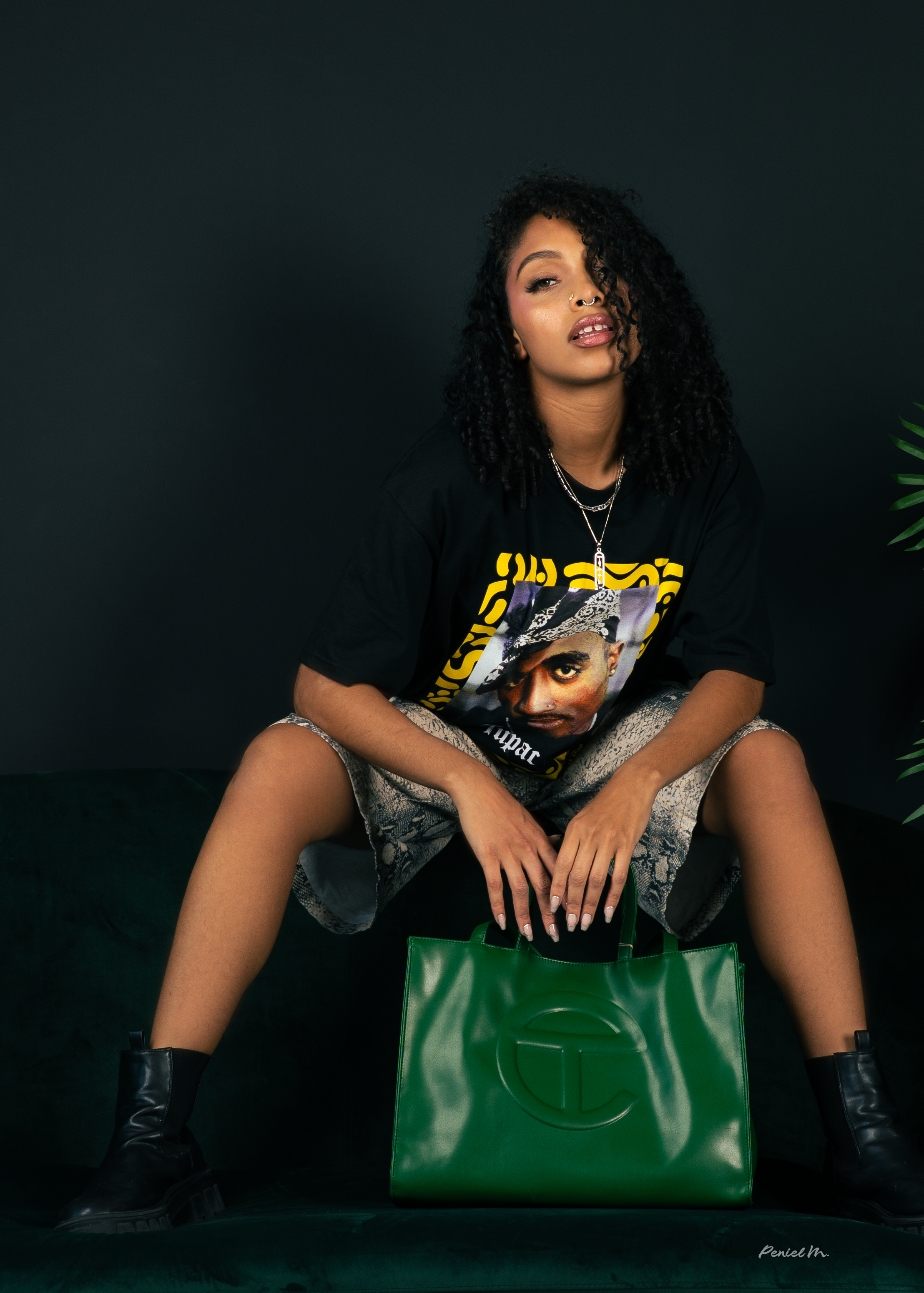 A young woman with curly black hair is sitting on a dark green velvet couch against a black background. She is wearing a black T-shirt with a printed face and yellow text, patterned shorts, black boots, and silver jewelry. She is holding a large gree