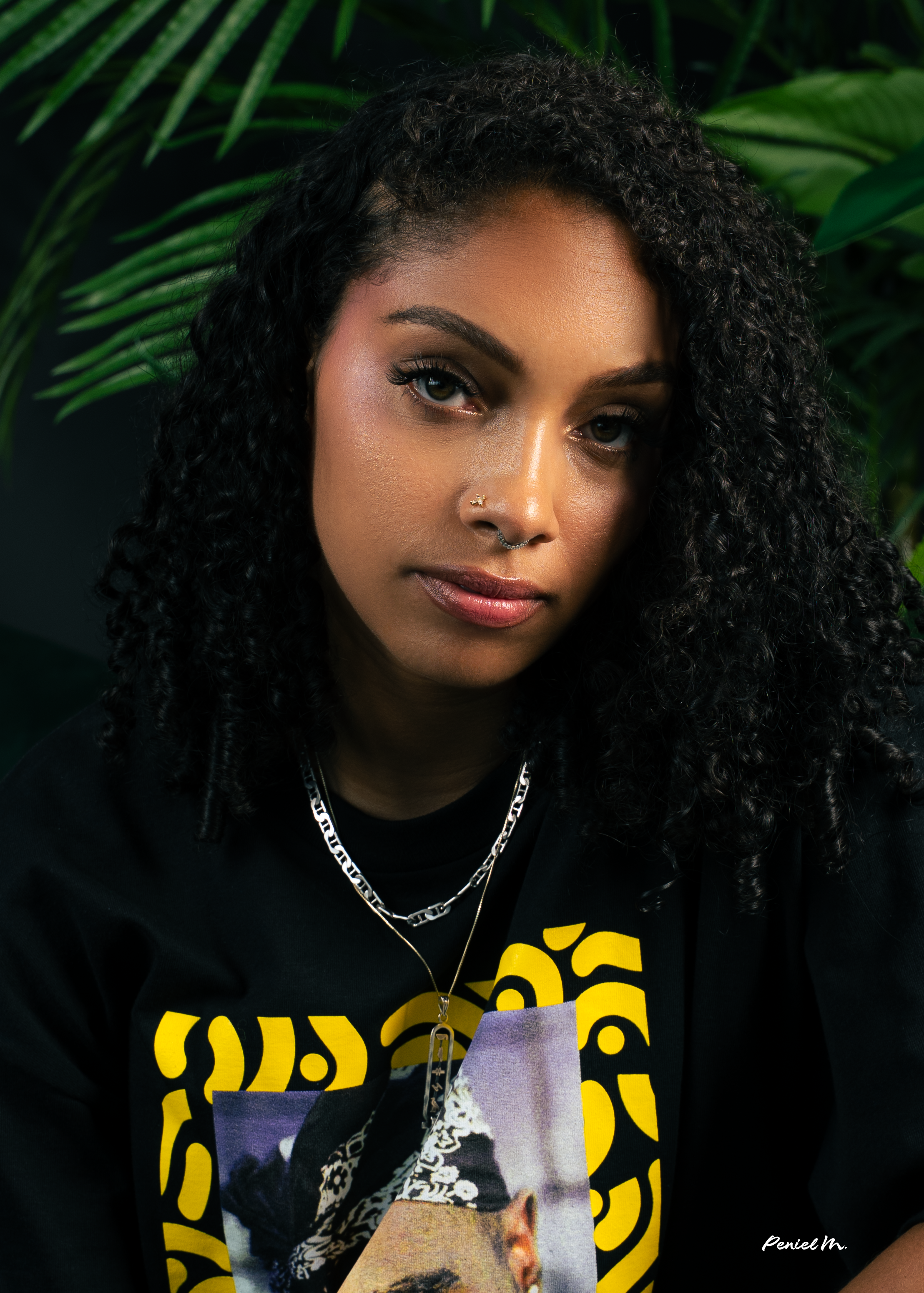 A young woman with dark curly hair, wearing layered necklaces, nose piercings, and a black graphic T-shirt, is posing in front of green tropical plants.