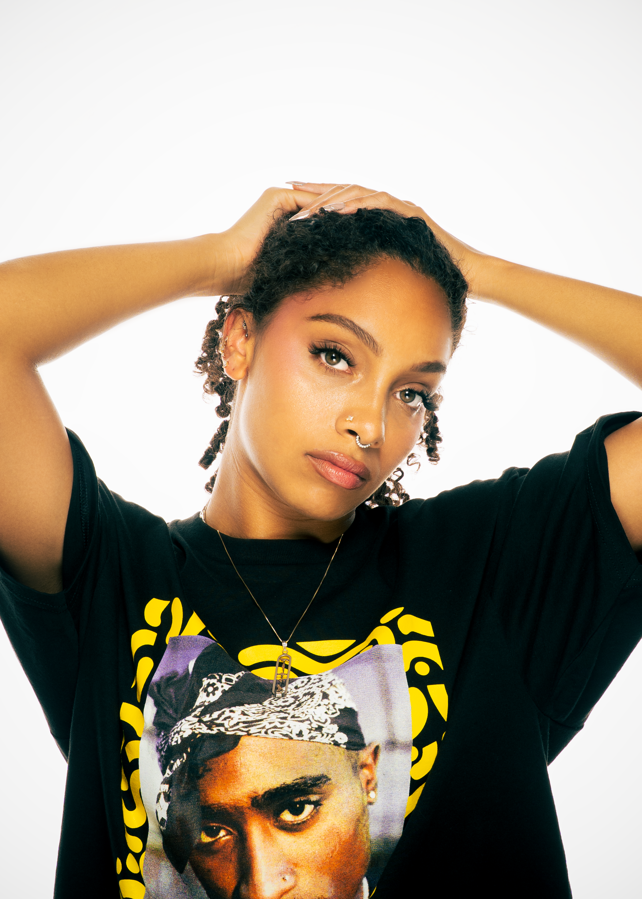 A woman with medium brown skin, curly hair, and blue-green eyes is posing with her arms raised behind her head. She is wearing a black T-shirt with a graphic print of a famous portrait of Tupac Shakur, a necklace with a key pendant, and has facial pi