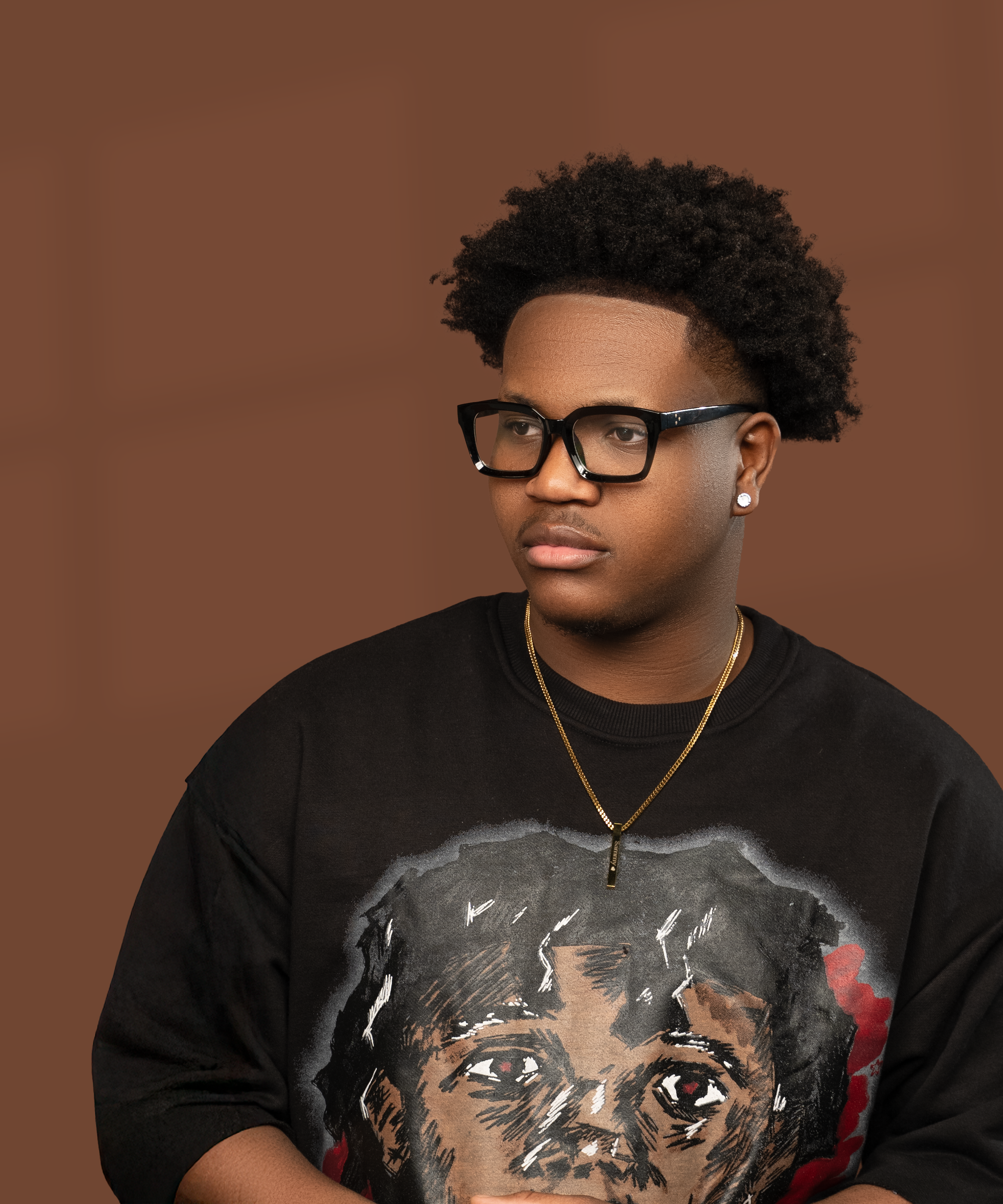 Young man with glasses, earrings, and a gold necklace, wearing a black sweatshirt with a graphic of a child's portrait, standing against a brown background.
