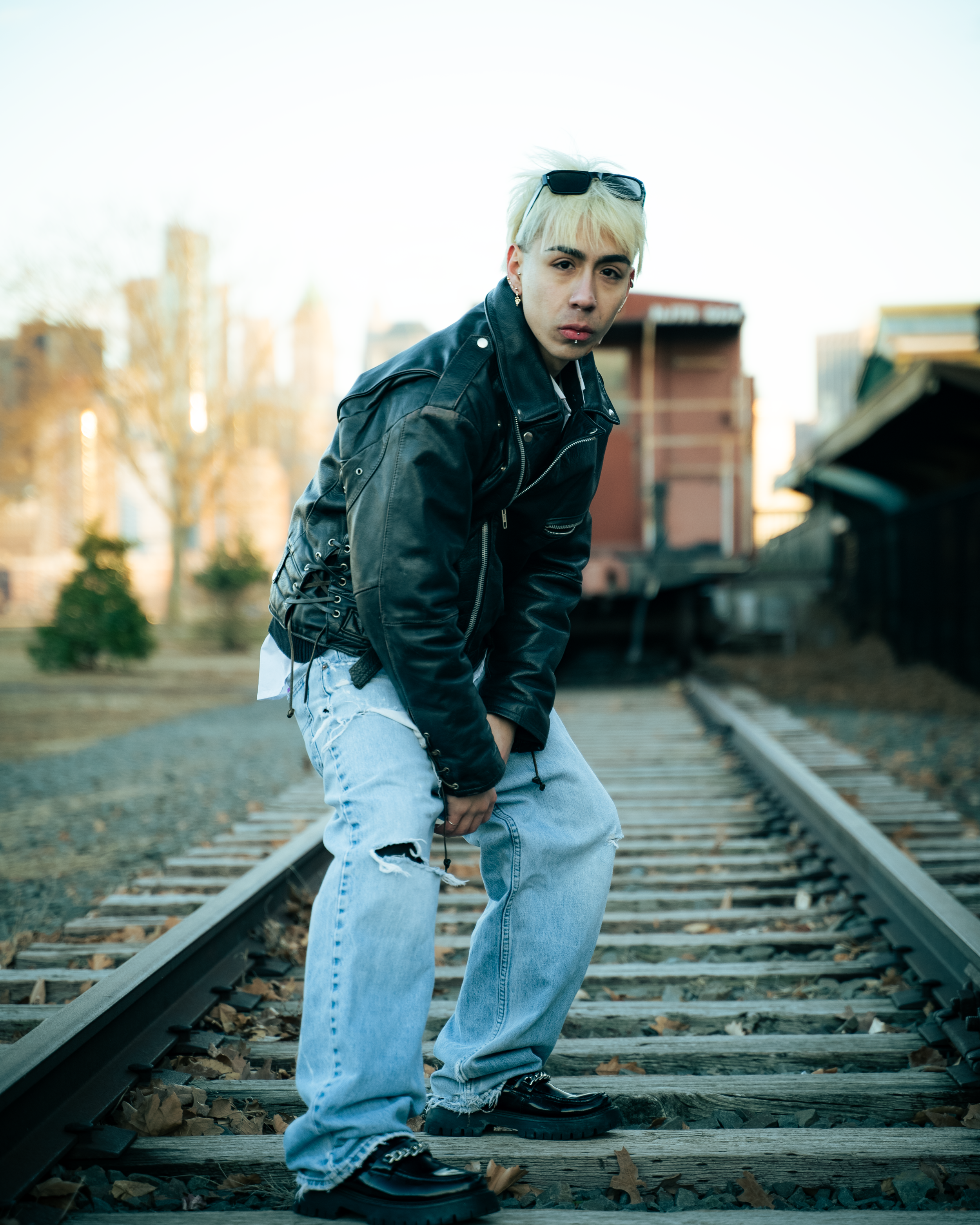 Person with bleached blonde hair, wearing a black leather jacket, ripped jeans, and sunglasses on their head, standing on train tracks with urban background.