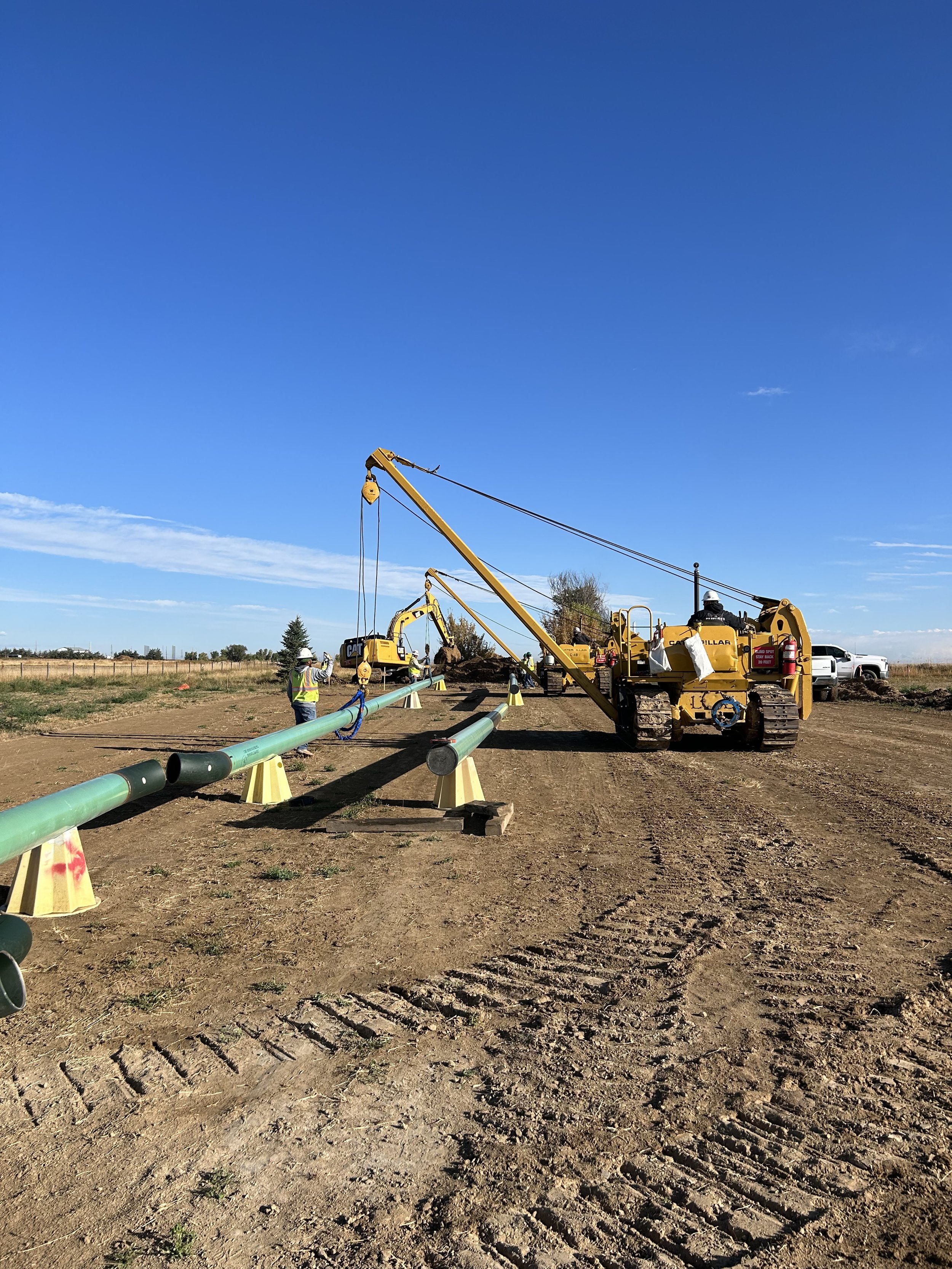 Construction workers and machinery installing a large pipeline in an open area with clear blue skies.
