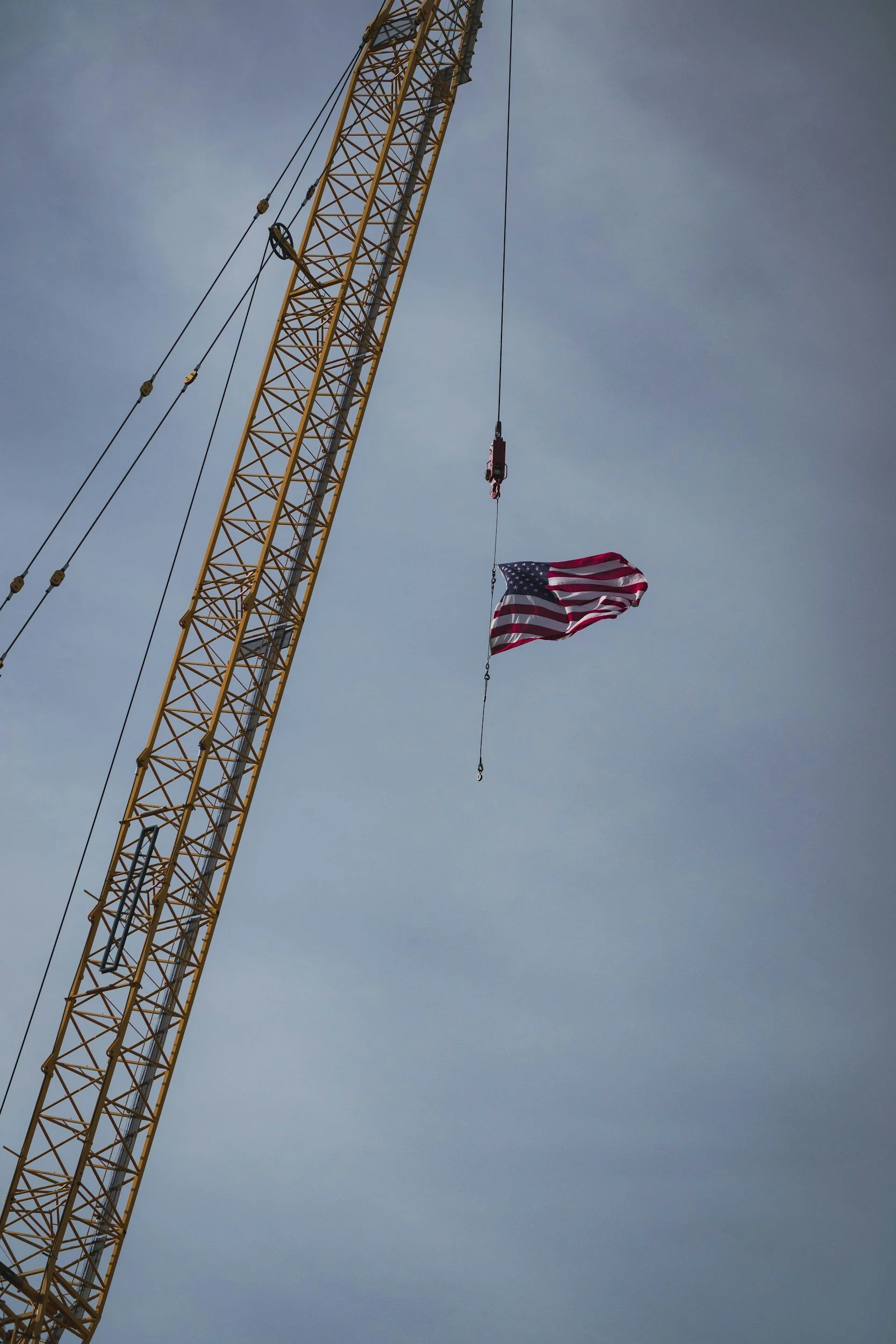 A construction crane with a flag of the United States hanging from it against a cloudy sky.