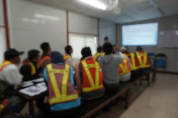 A group of people sitting at a long table in a classroom, wearing yellow and red safety vests, facing a presenter and a large screen or projector at the front of the room.