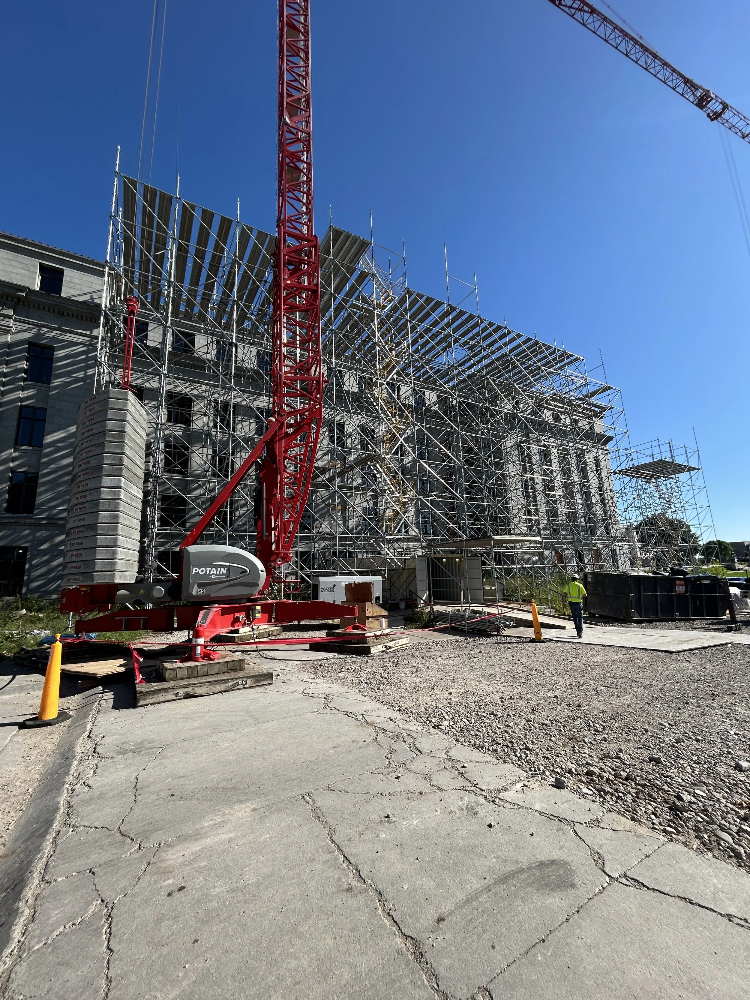 Construction site with scaffolding around a historic building, large red tower crane, and a worker in a yellow safety vest.