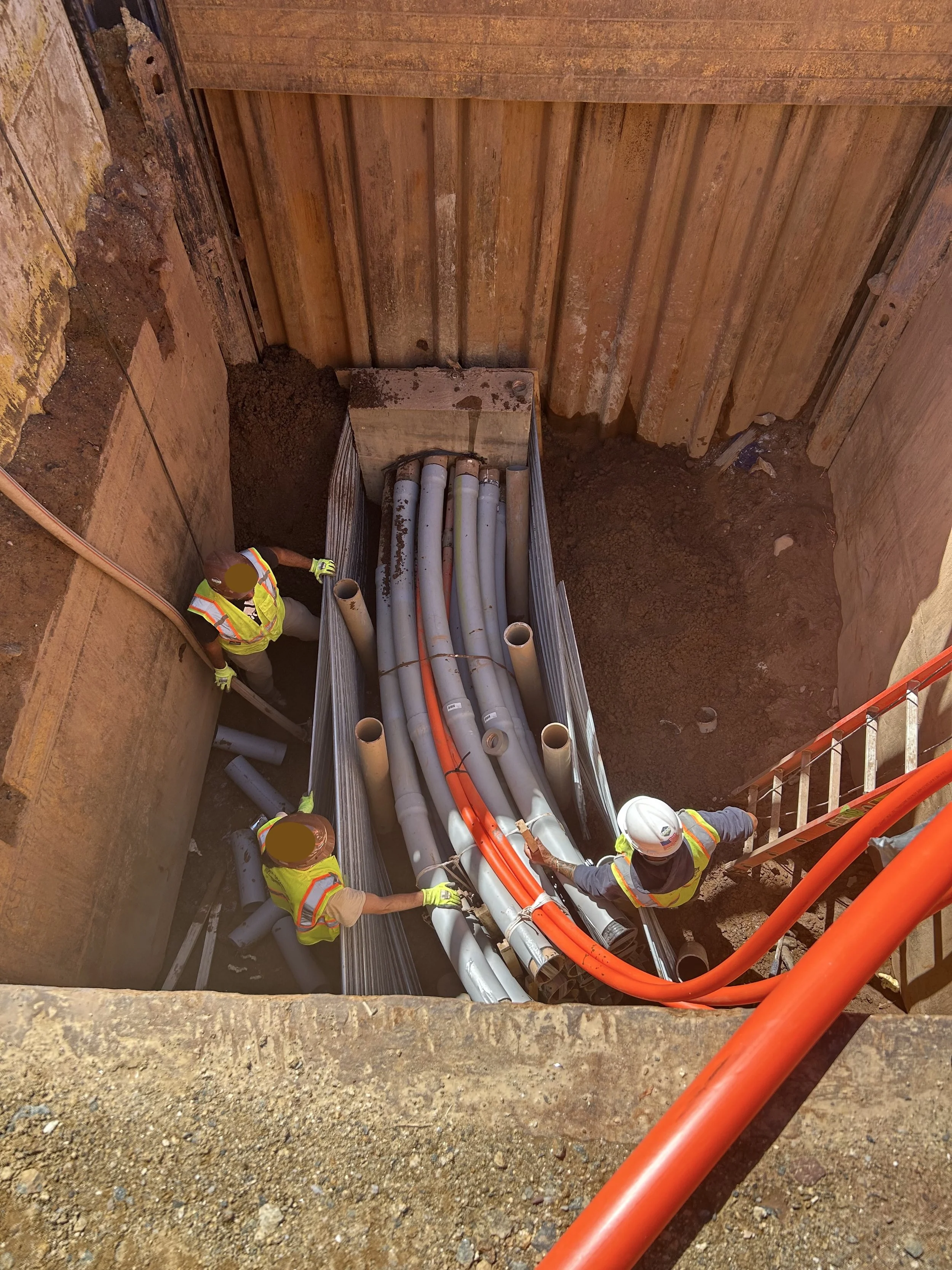 Three construction workers working on underground piping installation in a deep excavation. The workers are wearing safety vests and helmets, and are handling large gray and orange pipes.
