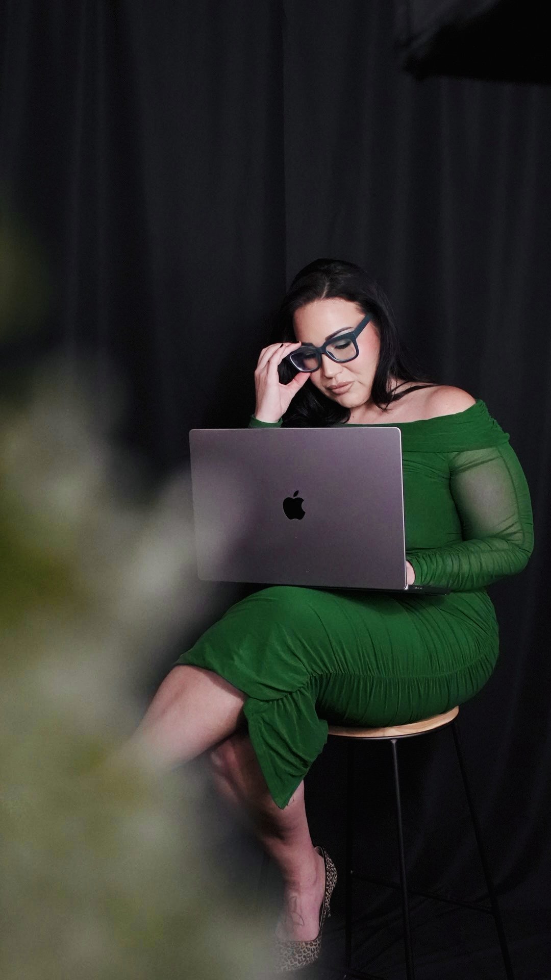 A woman with black hair and glasses wearing a green dress, sitting on a stool, working on a silver MacBook against a black curtain background