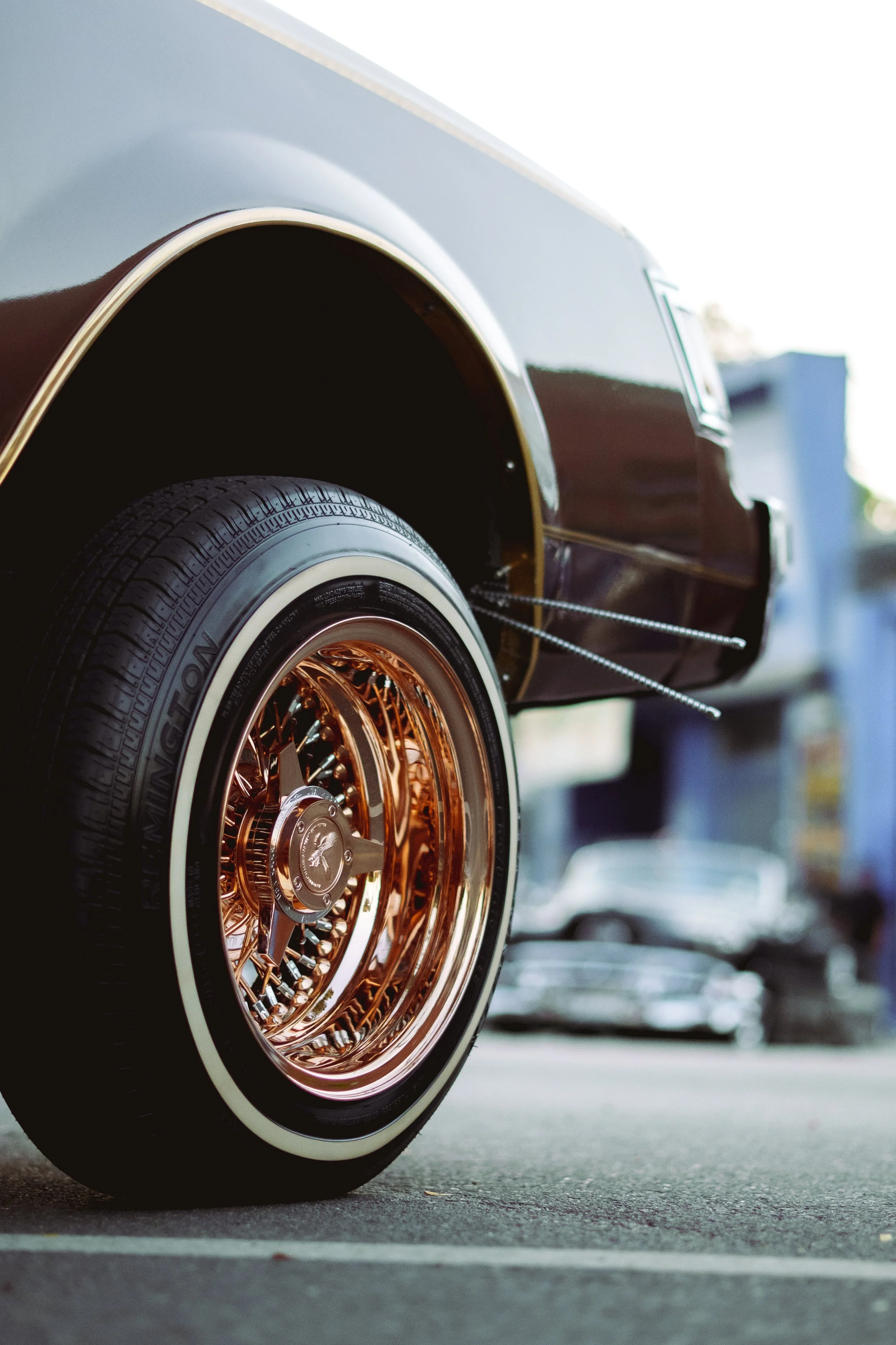 Close-up of a vintage car's wheel with a gold wire spoke rim and a white wall tire, parked outdoors.