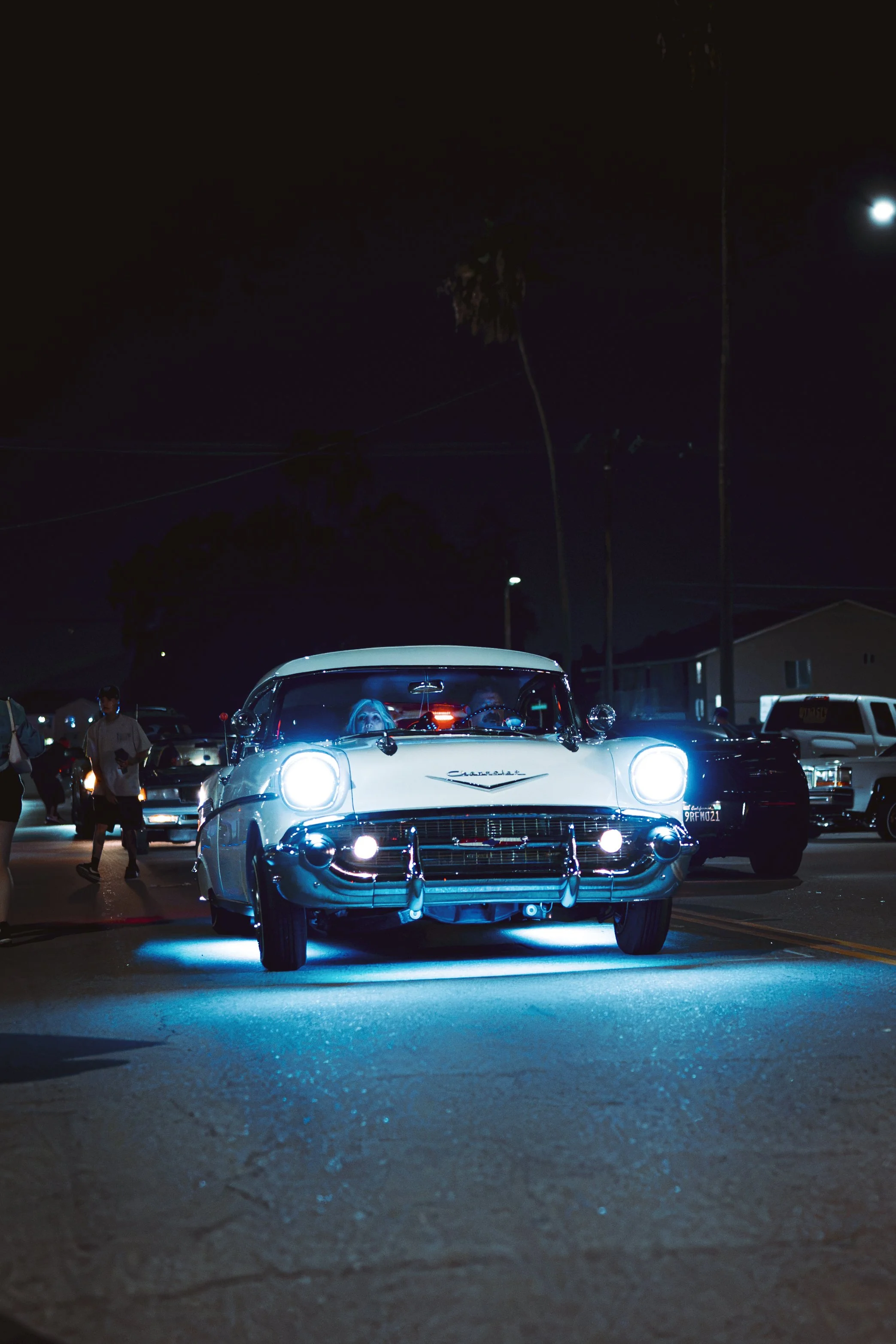 A nighttime scene of a classic white Chevrolet Corvette parked in a lot, illuminated by blue underglow lights. People are walking around, and there are other cars and houses visible in the background, with the moon shining in the dark sky.