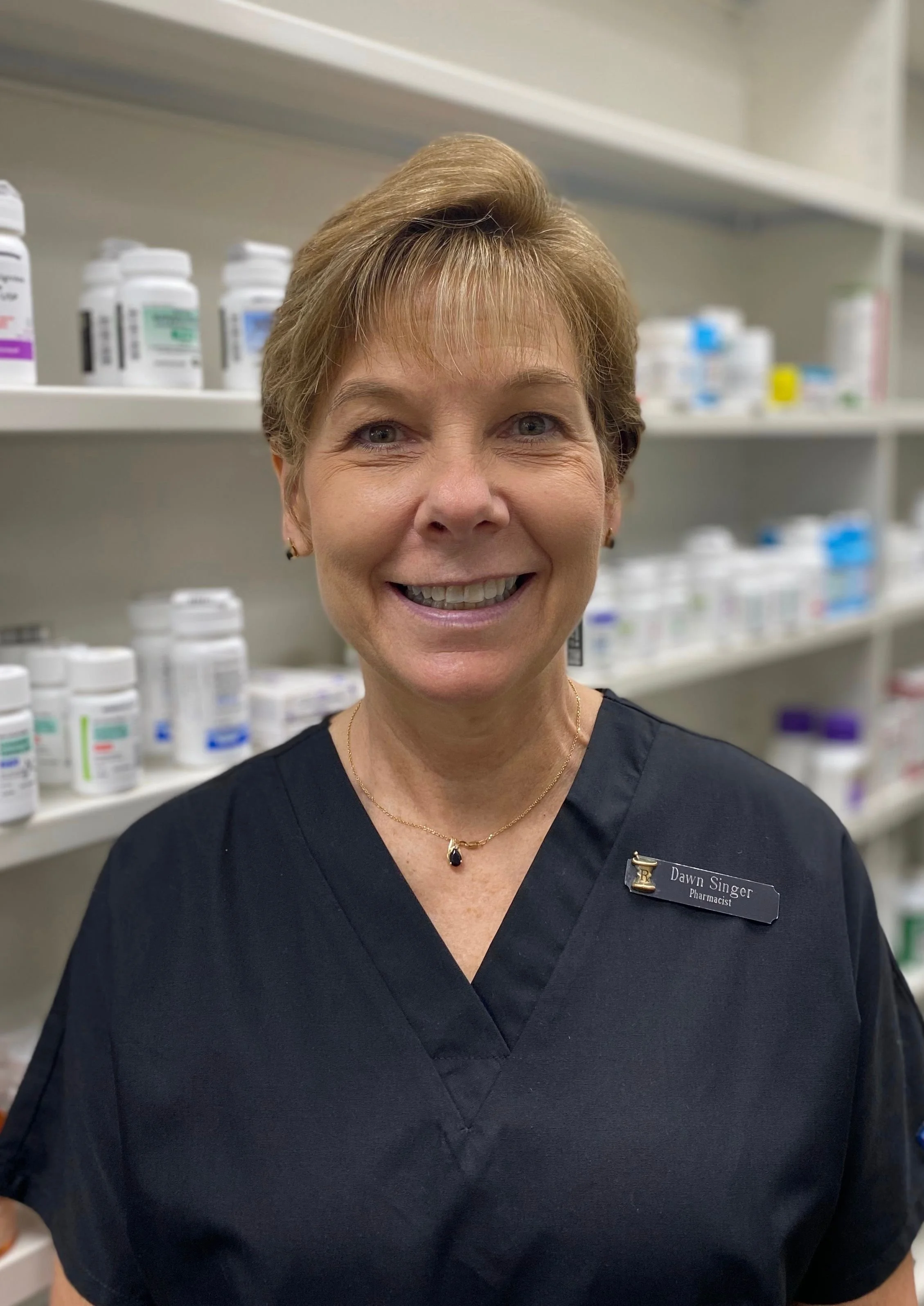 A smiling woman wearing a black medical uniform with a name tag that reads Dawn Singer, standing in front of pharmacy shelves filled with bottles and medication containers.