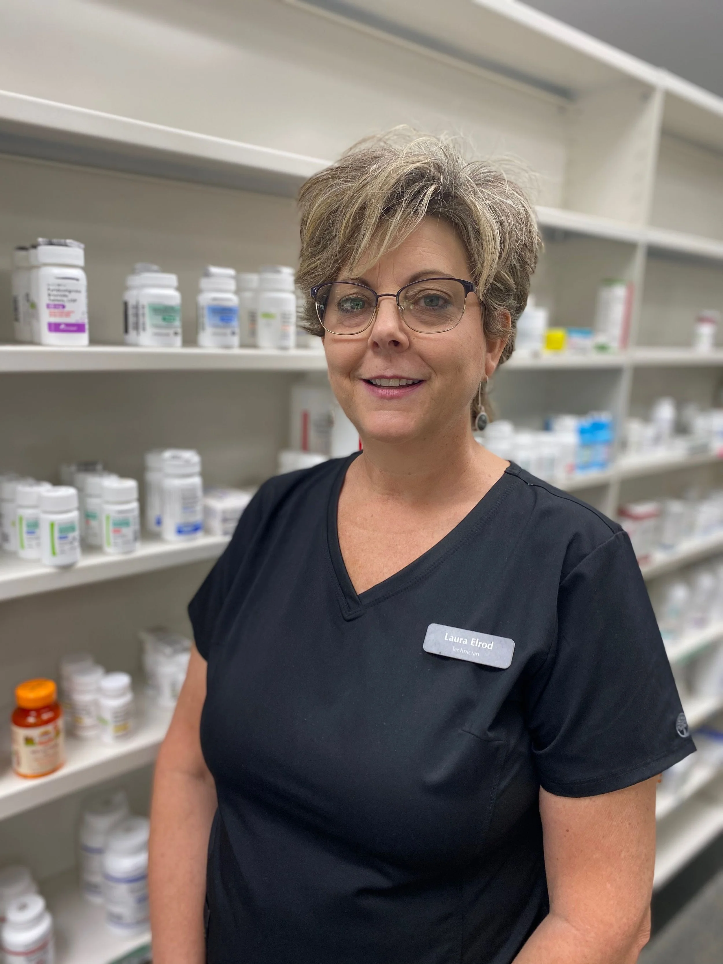 A woman with short gray hair, glasses, and a black uniform with a name tag that reads Laura Elrod, standing in front of shelves stocked with various bottles of medication or supplements.