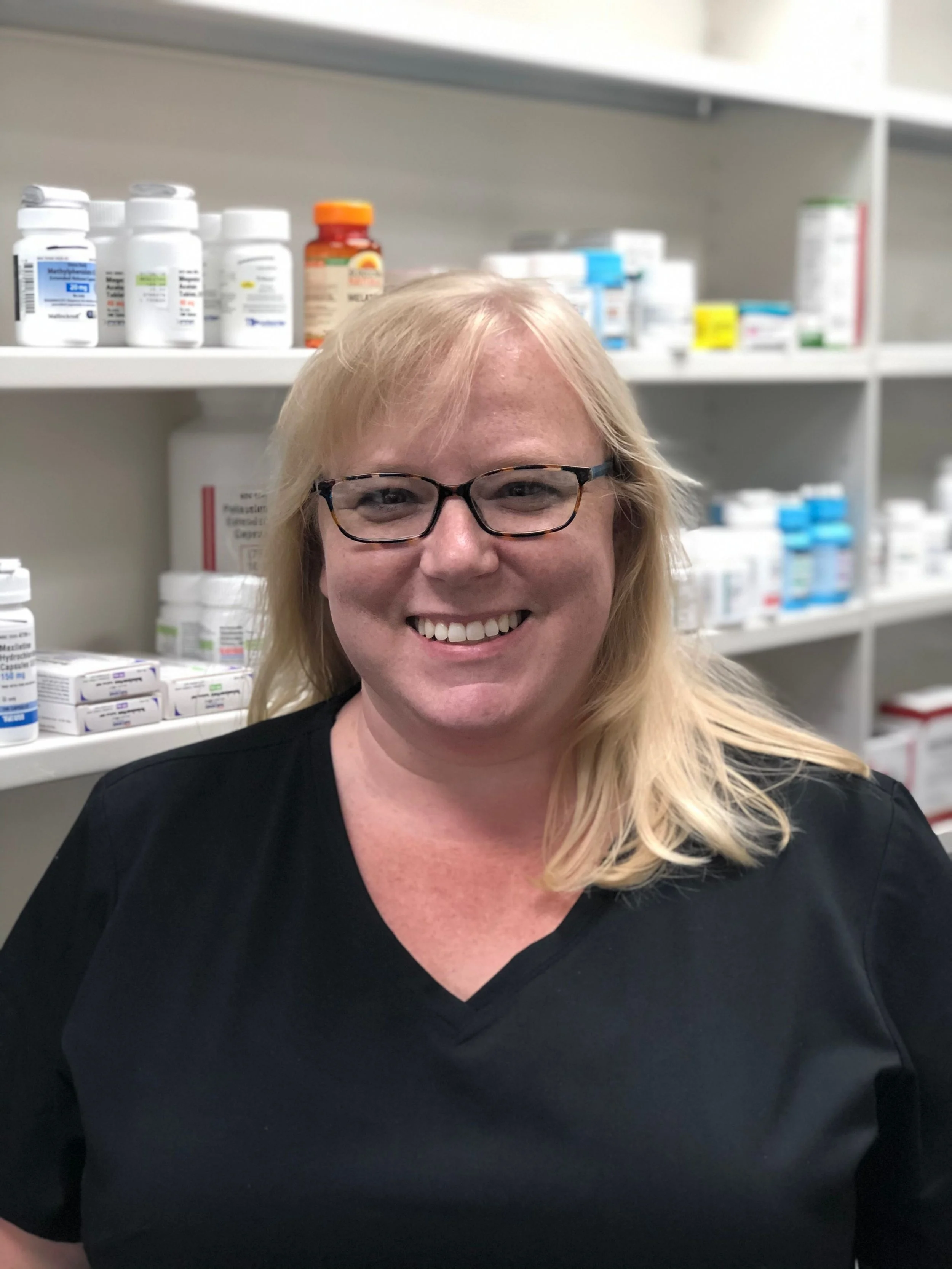 A smiling woman with blonde hair and glasses, wearing a black scrub top, sitting in front of pharmacy shelves filled with prescription bottles and medication boxes.