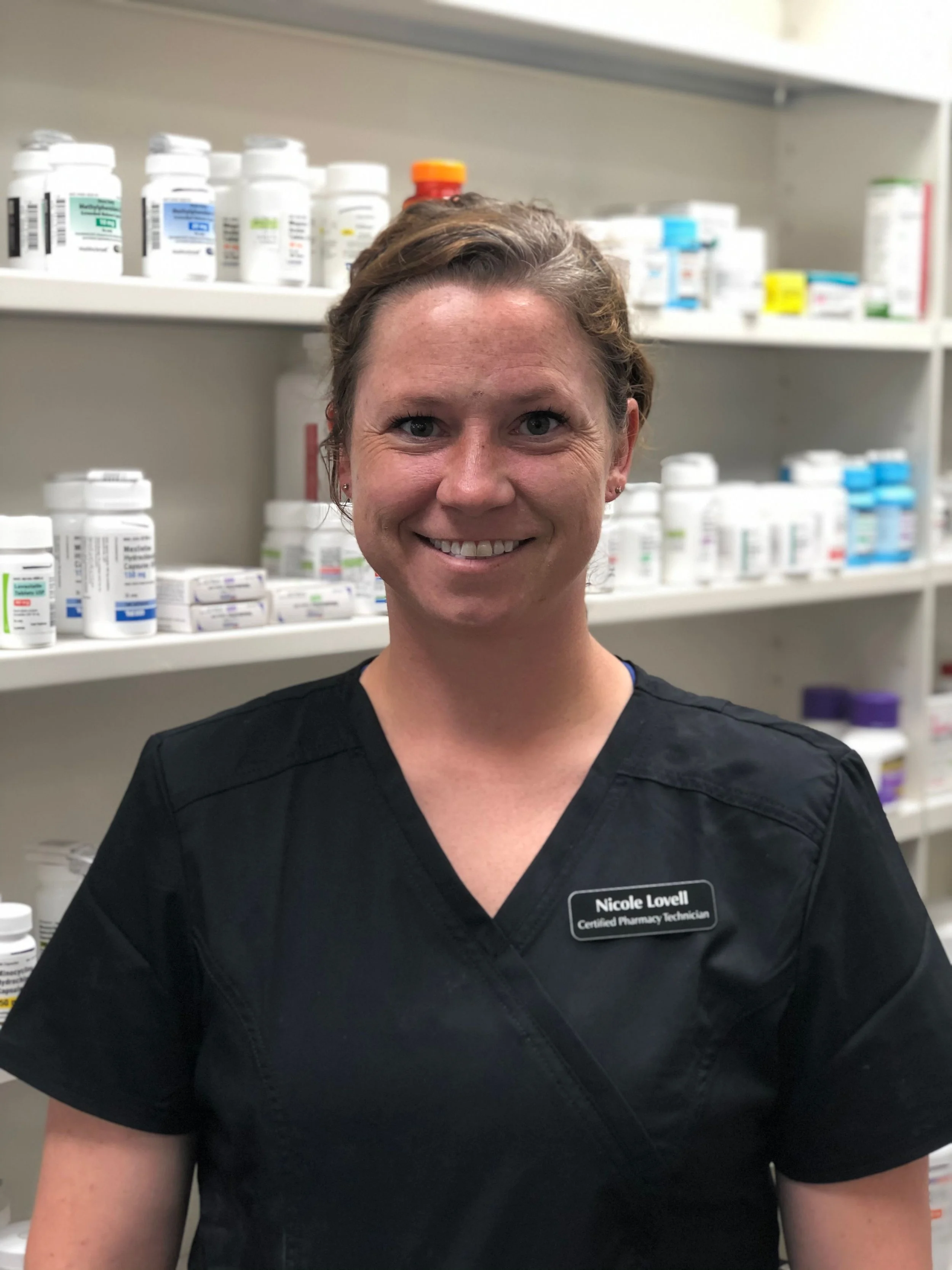 A woman smiling in a pharmacy, wearing a black uniform with a name tag that reads 'Nicole Lowell, Certified Pharmacy Technician,' with shelves of medication bottles in the background.