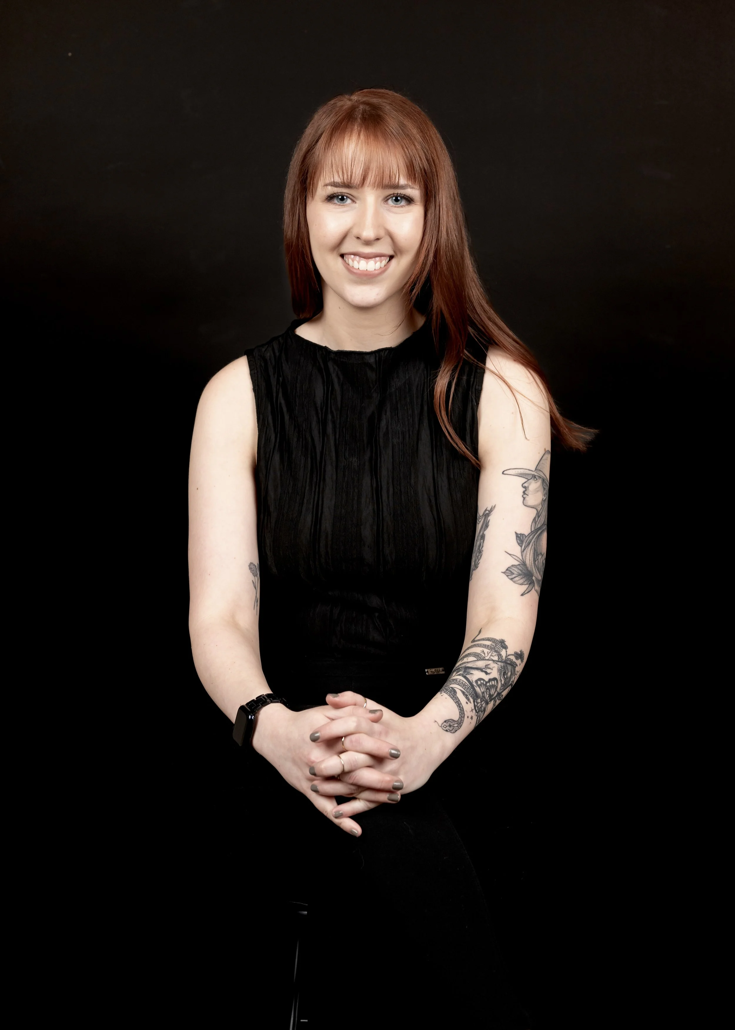 A photo of trauma therapist and counselor, Sydney Strother, LPCA, in Aiken, SC, that offers therapy for depression and anxiety. She is a woman with red hair and tattoos, smiling, wearing a black sleeveless top, against a black background.