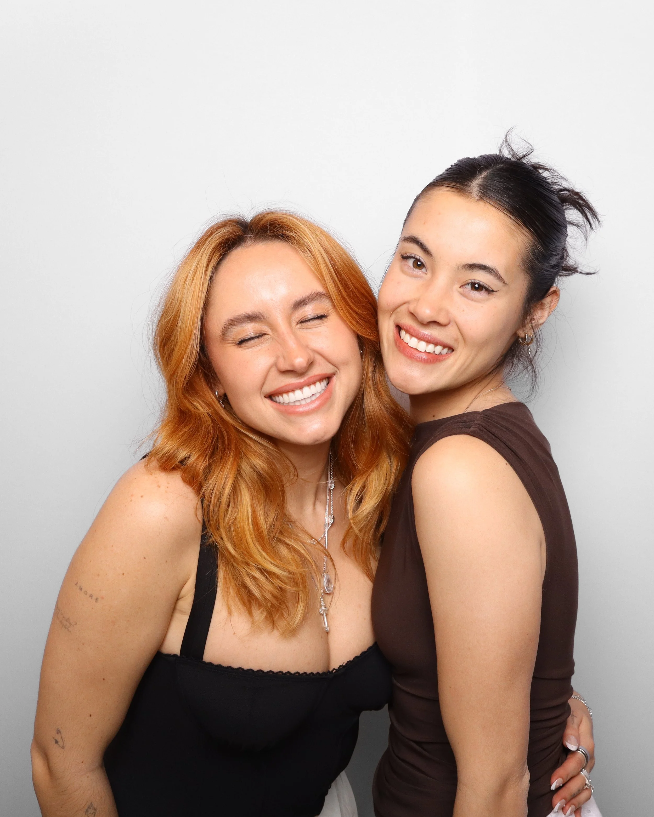 Two smiling women with their heads close together, one with red hair and the other with dark hair, standing against a plain light gray background.