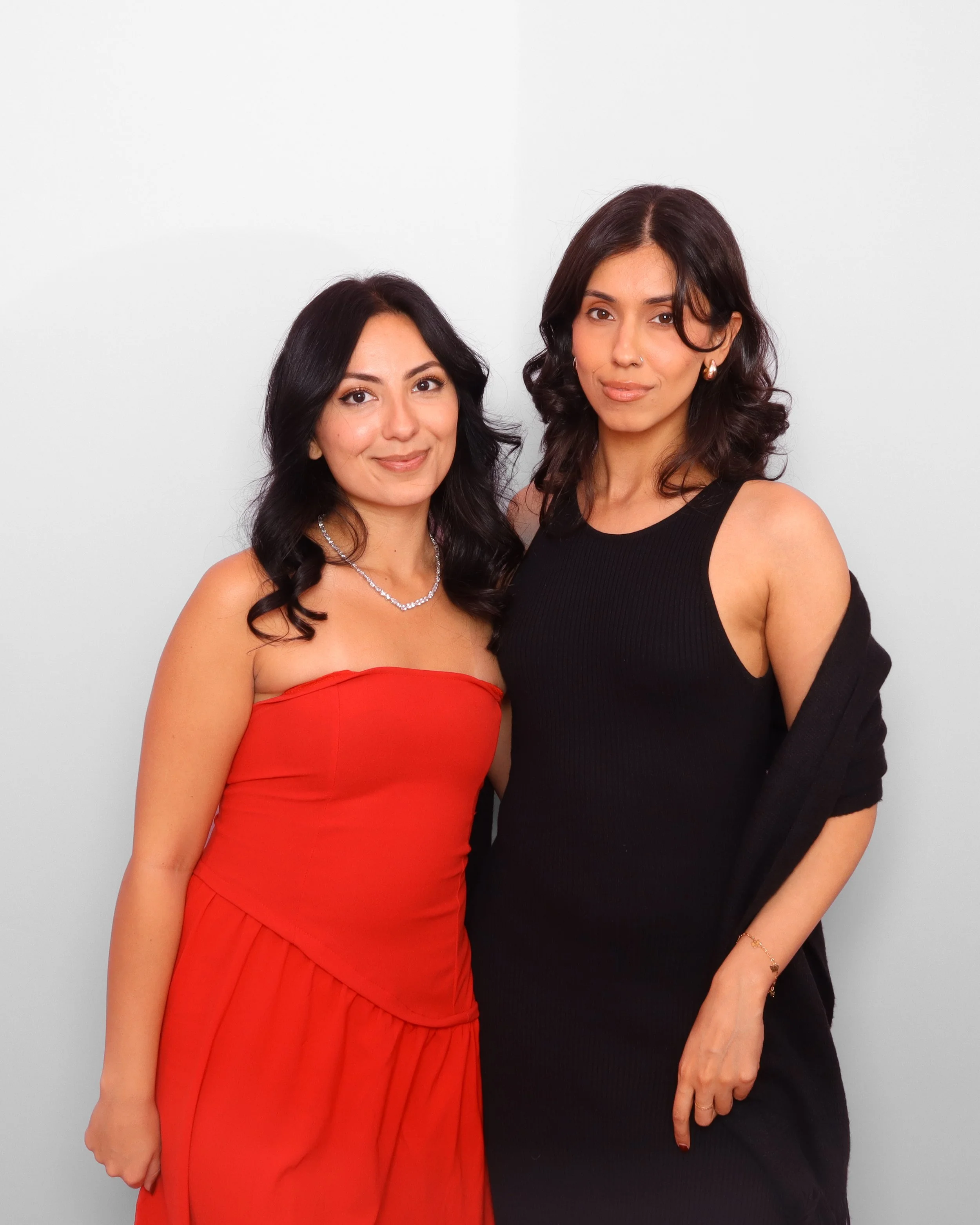 Two women standing side by side against a plain white background, smiling and looking at the camera. One is wearing a red strapless dress and the other a black top with an off-shoulder sleeve.