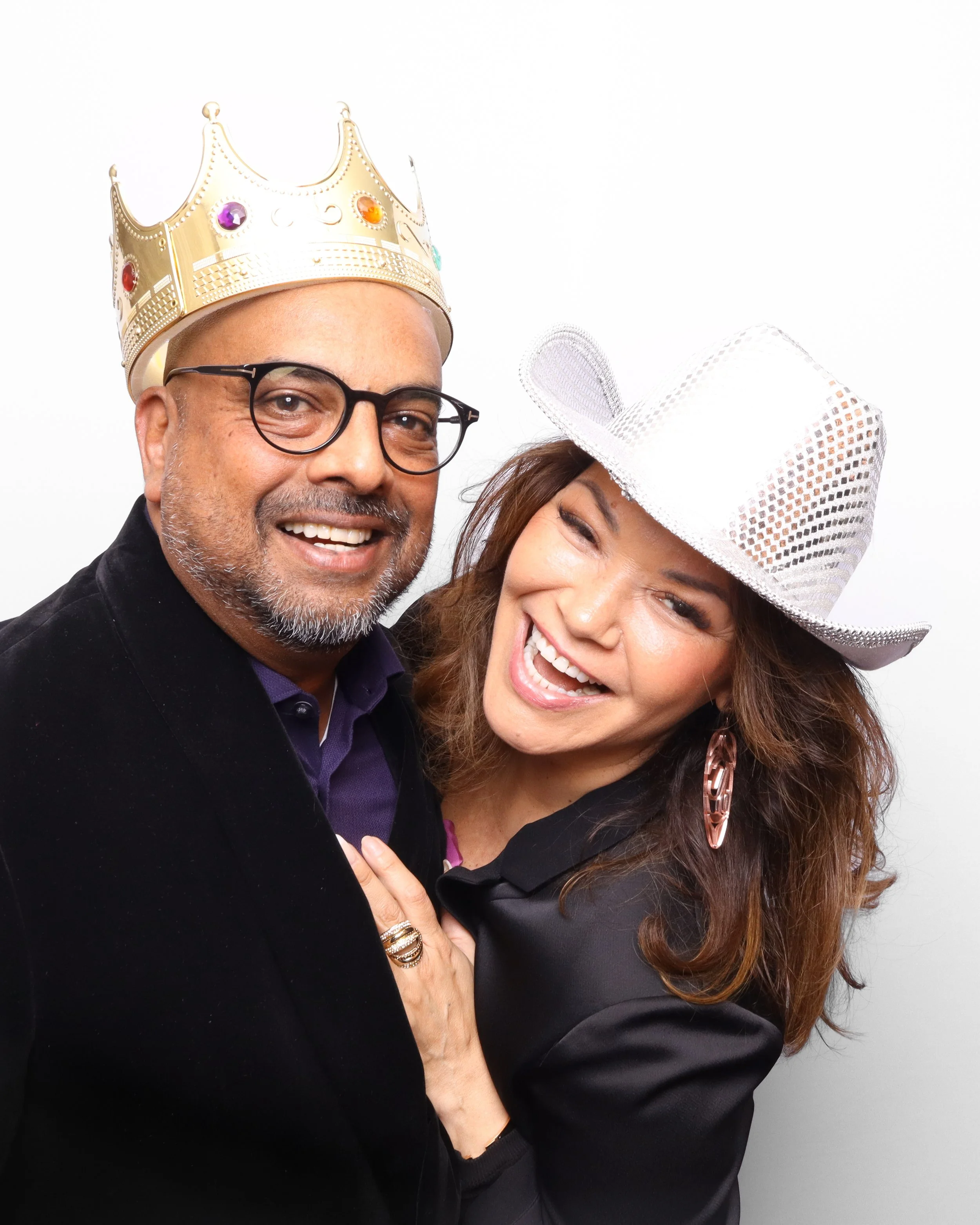 Close-up of a smiling man wearing a gold crown with colorful jewels and glasses, and a woman with a large white sparkly hat, smiling and showing her teeth.