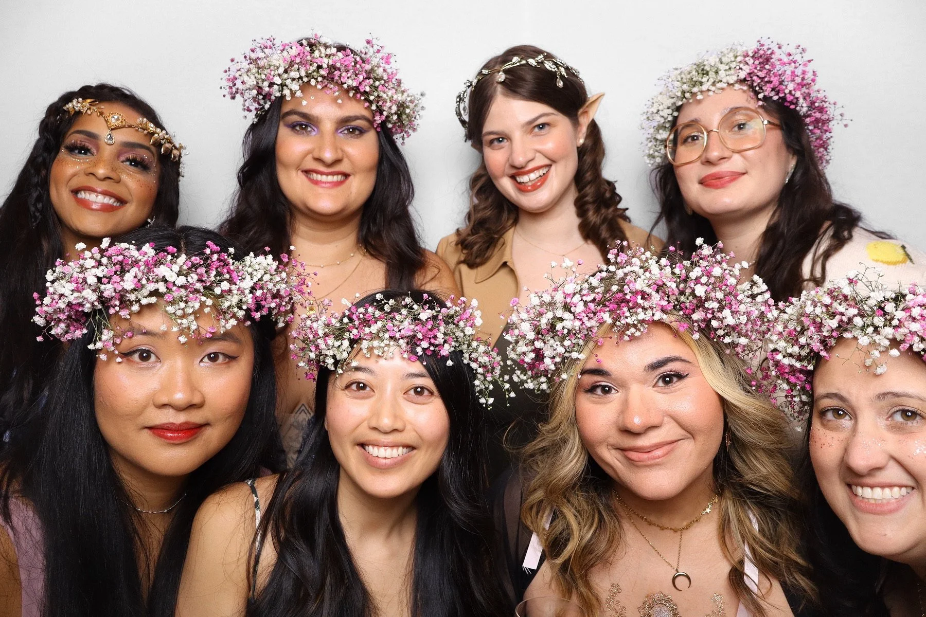 Group of women smiling, wearing floral crowns made of pink and white flowers, some with elf ears and glitter makeup, posing for a photo on a white background.
