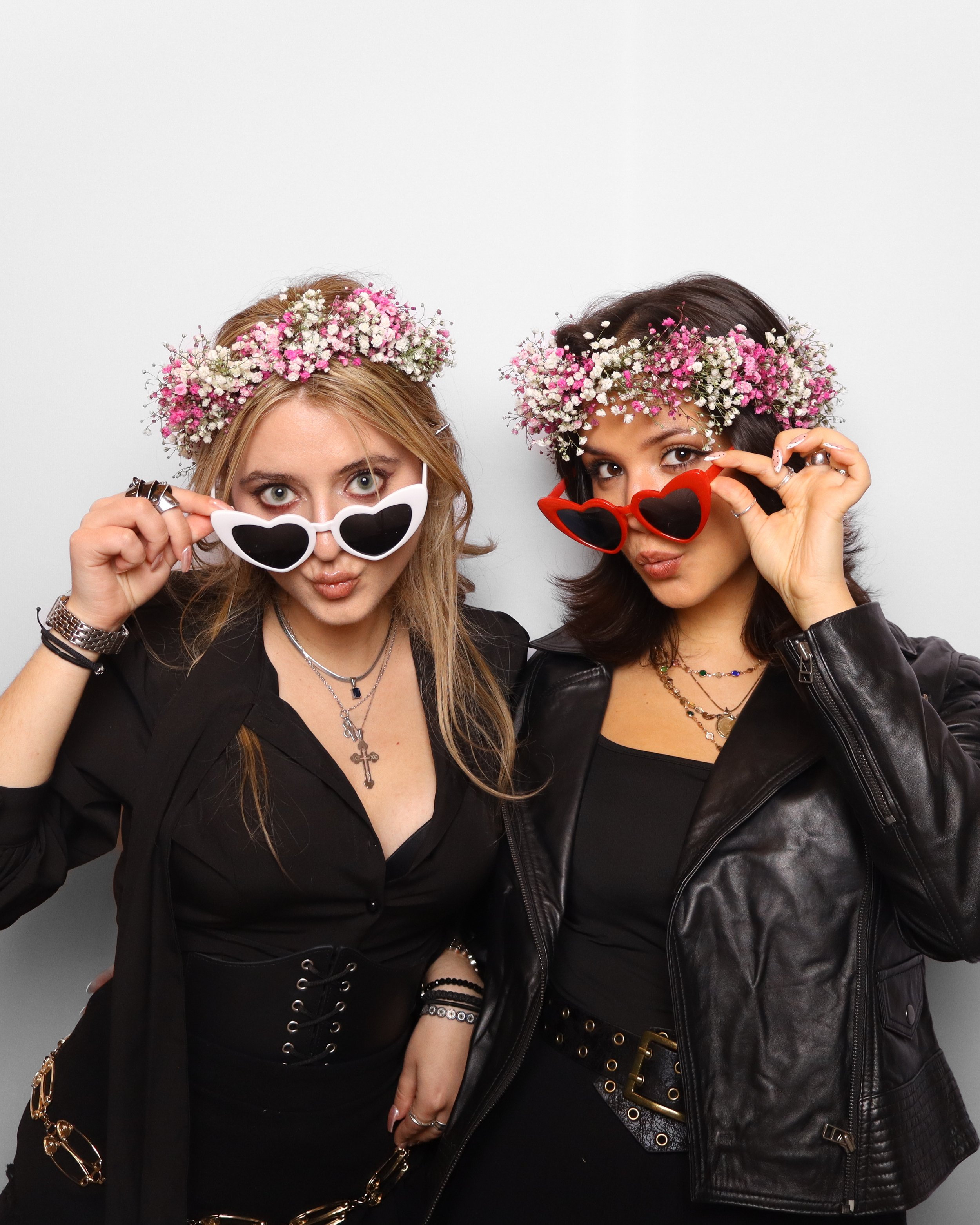 Two women wearing flower crowns and sunglasses posing for a photo against a plain background.