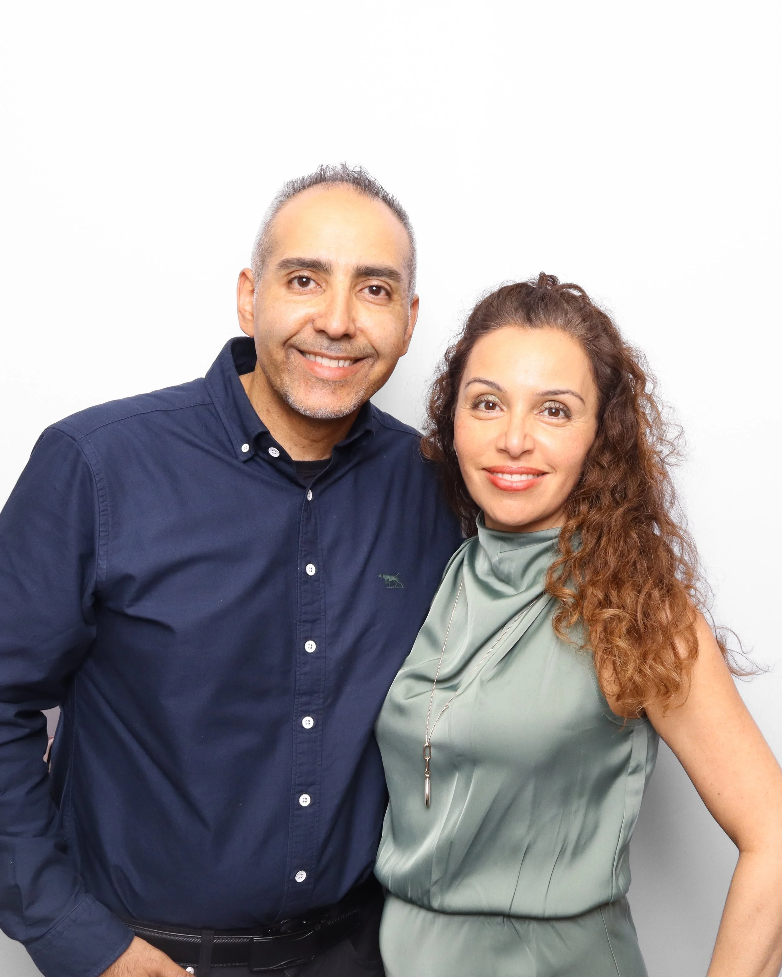 A man and woman smiling, standing close together in front of a plain white wall. The man is wearing a dark blue shirt, and the woman is wearing a light greenish-gray blouse with a necklace.