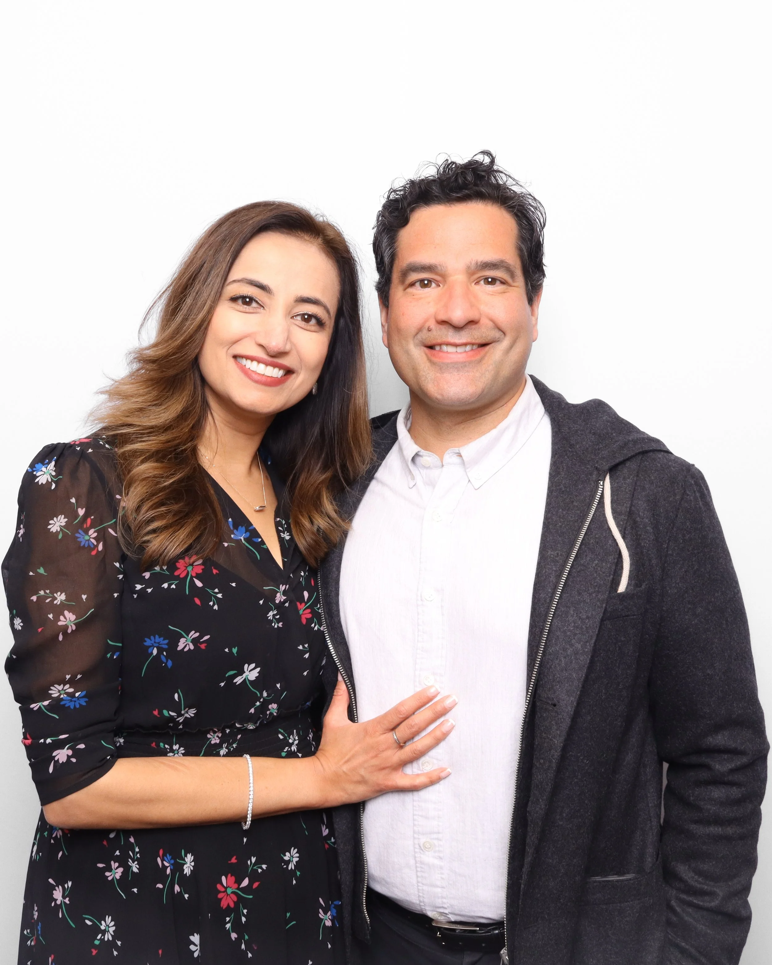 A woman with long brown hair wearing a black floral dress smiling and standing close to a man with black curly hair wearing a white shirt and a grey hoodie, both smiling against a plain white background.