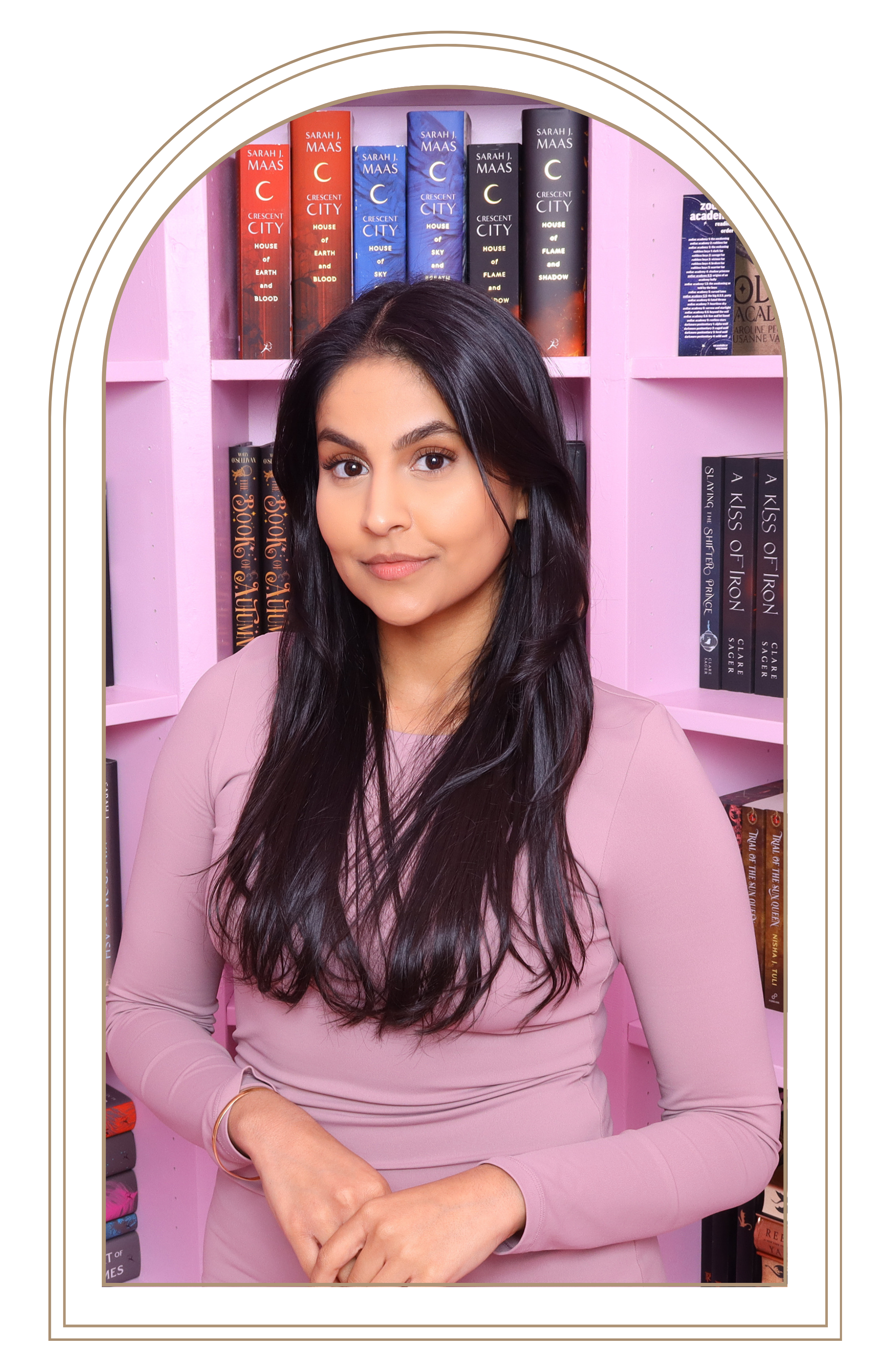 A woman with long dark hair, wearing a light pink long-sleeve shirt, standing in front of pink shelves filled with books.