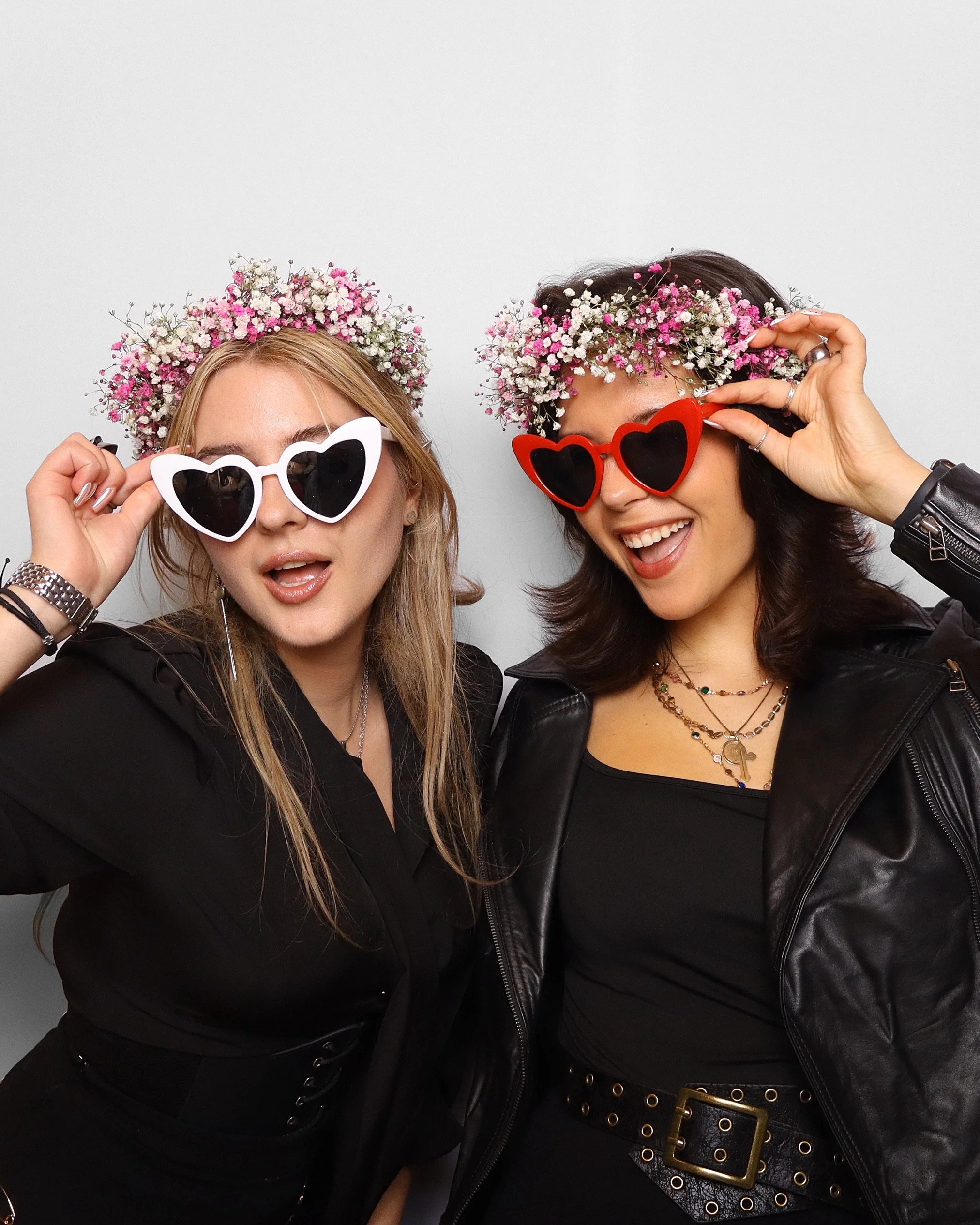 Two women wearing flower crowns and heart-shaped sunglasses, smiling and posing against a plain white background.