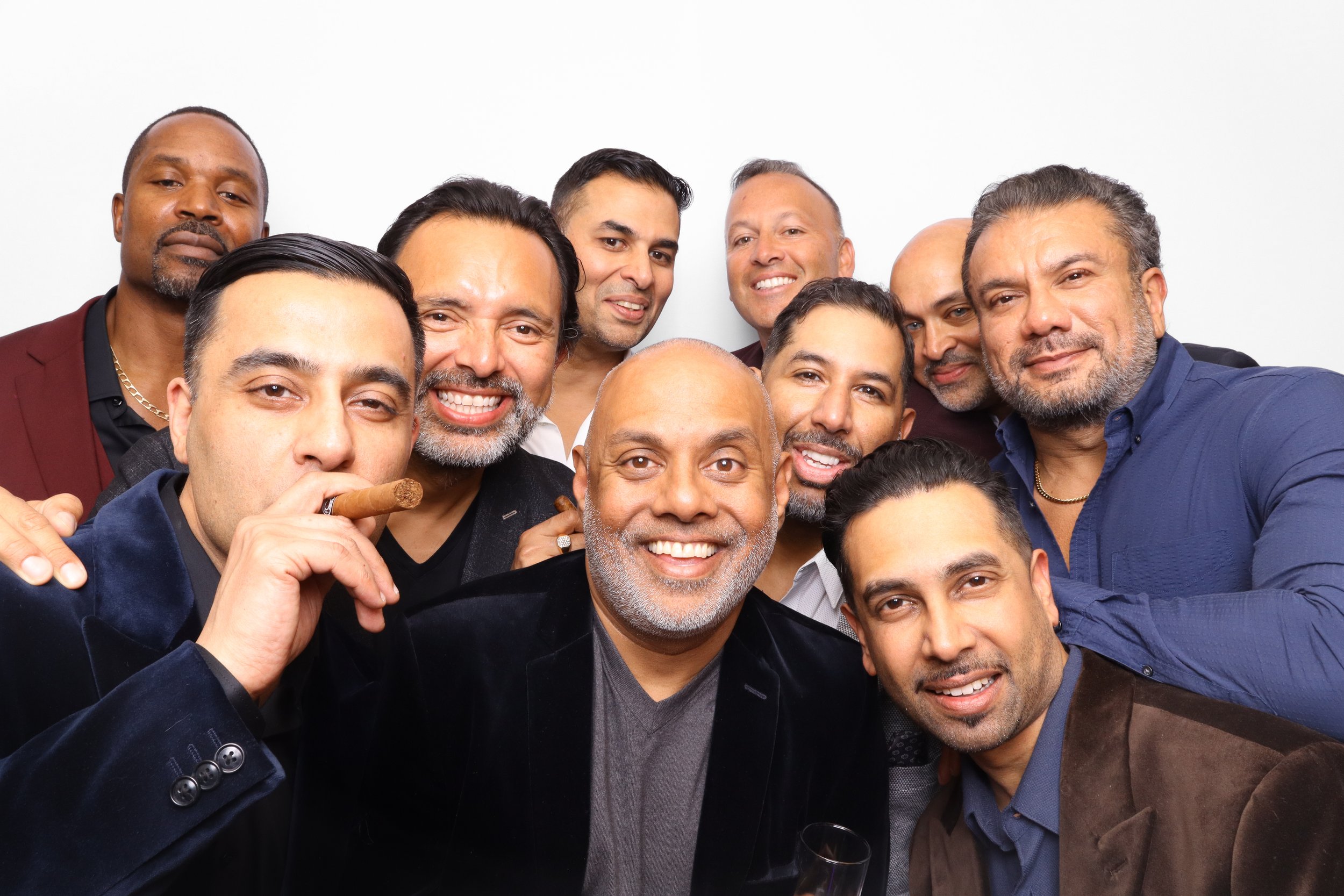 Group of diverse men smiling and taking a selfie together at a social event, some holding cigars and glasses, against a plain white background.