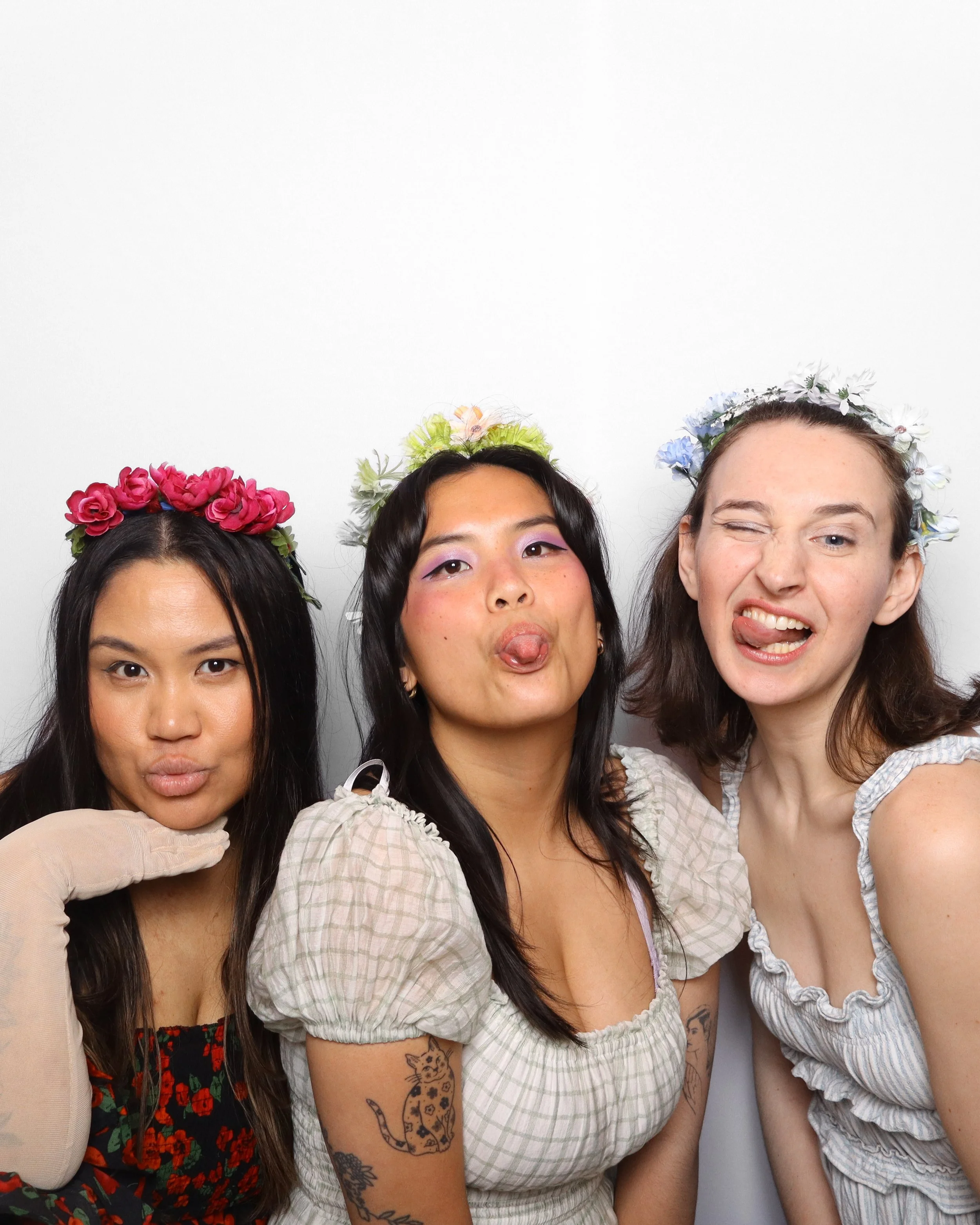 Three women wearing flower crowns and making funny faces in front of a plain white background.