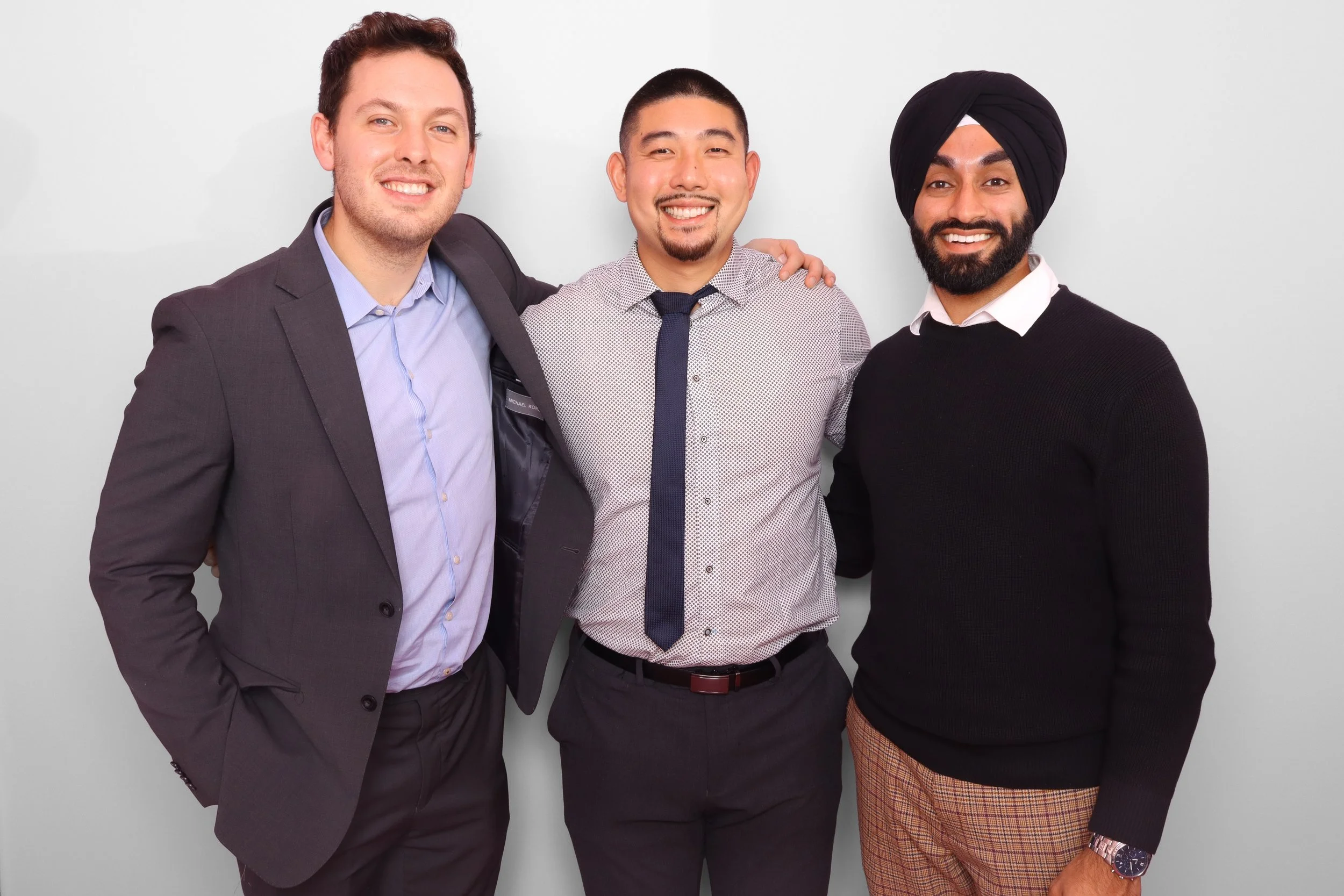 Three men standing together smiling, one wearing a suit, one in a patterned shirt and tie, and the third wearing a turban and a black sweater.