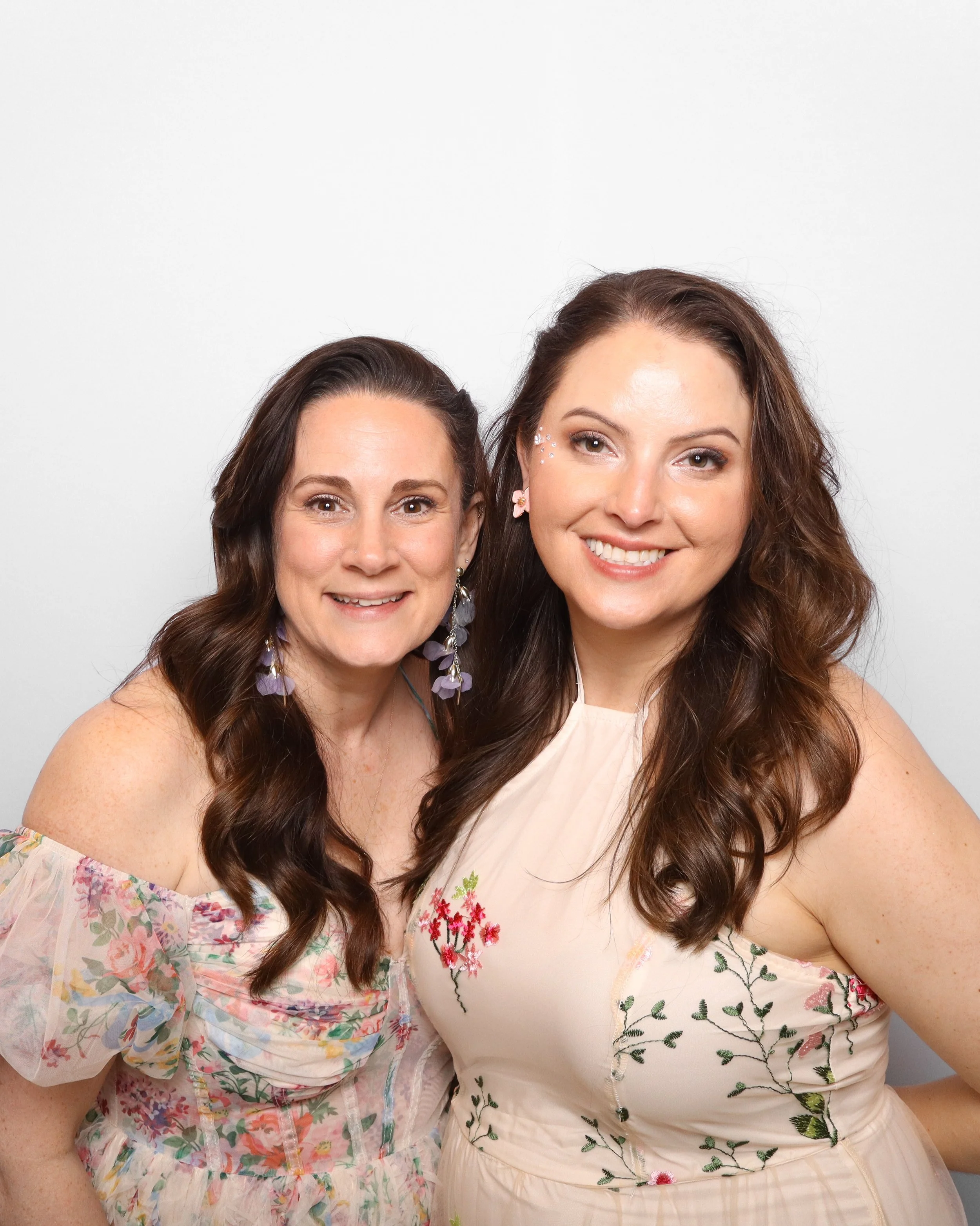 Two women smiling, wearing floral dresses, close-up portrait, plain gray background.
