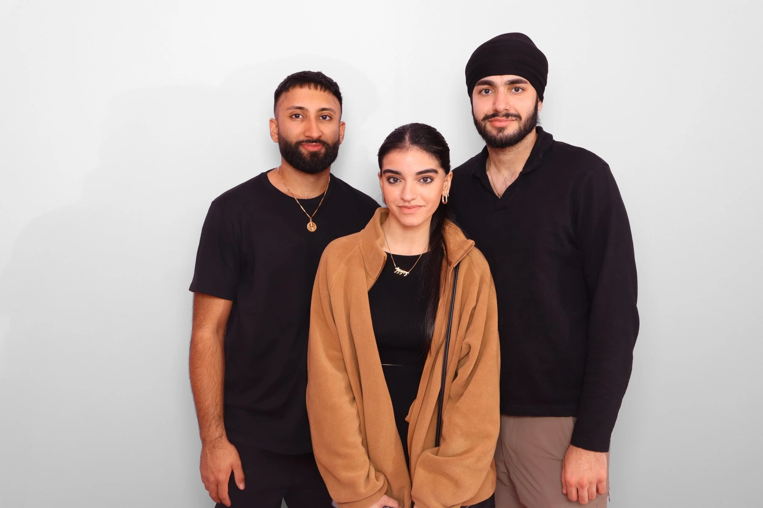 Three young adults standing together against a white background, looking at the camera. The group includes a woman in the center wearing a tan jacket, a man with a beard and black shirt to her left, and a man with a turban and black shirt to her righ