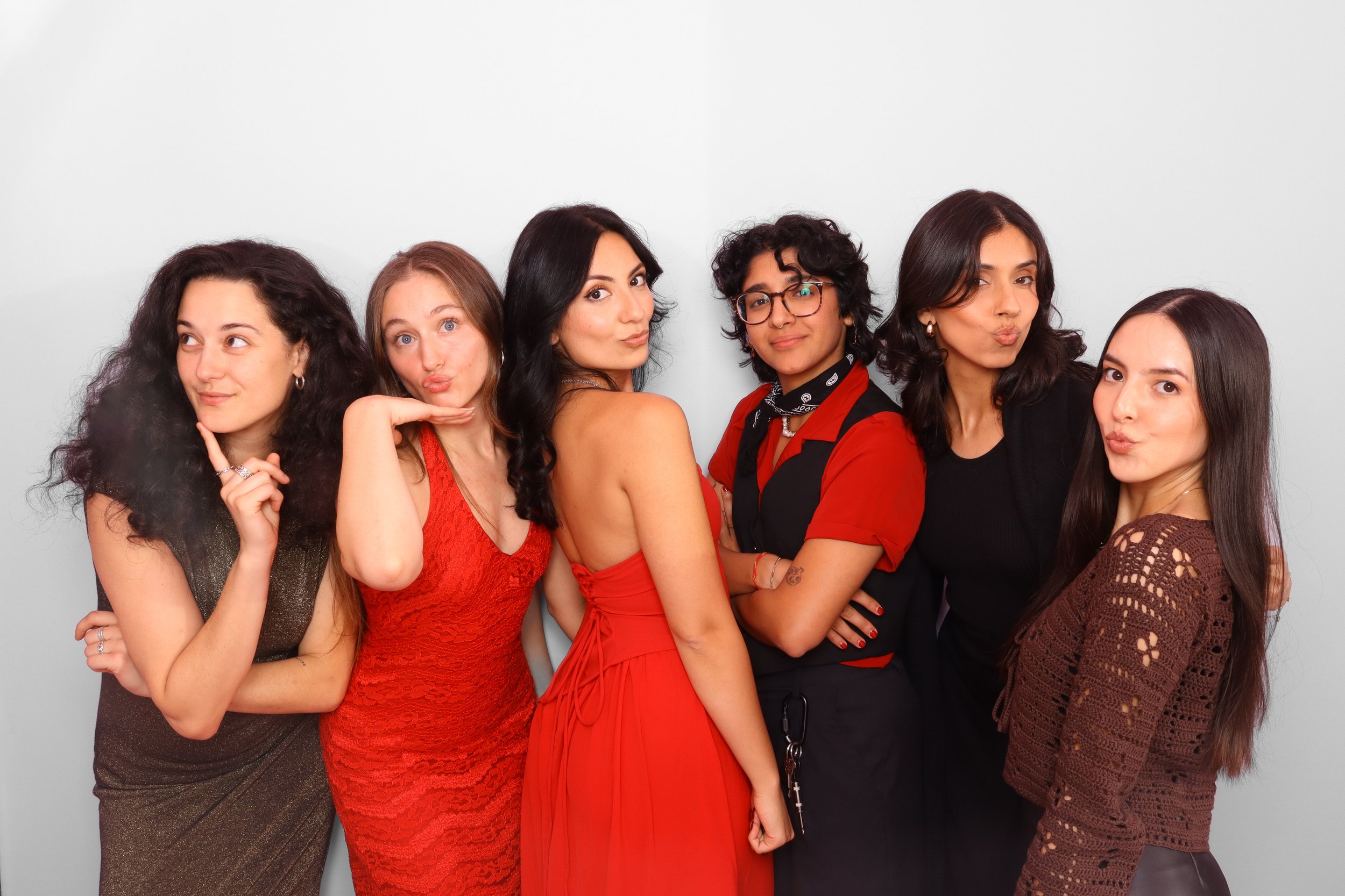 Group of six women posing together against a plain white background, with different hairstyles and outfits, making various facial expressions and gestures.