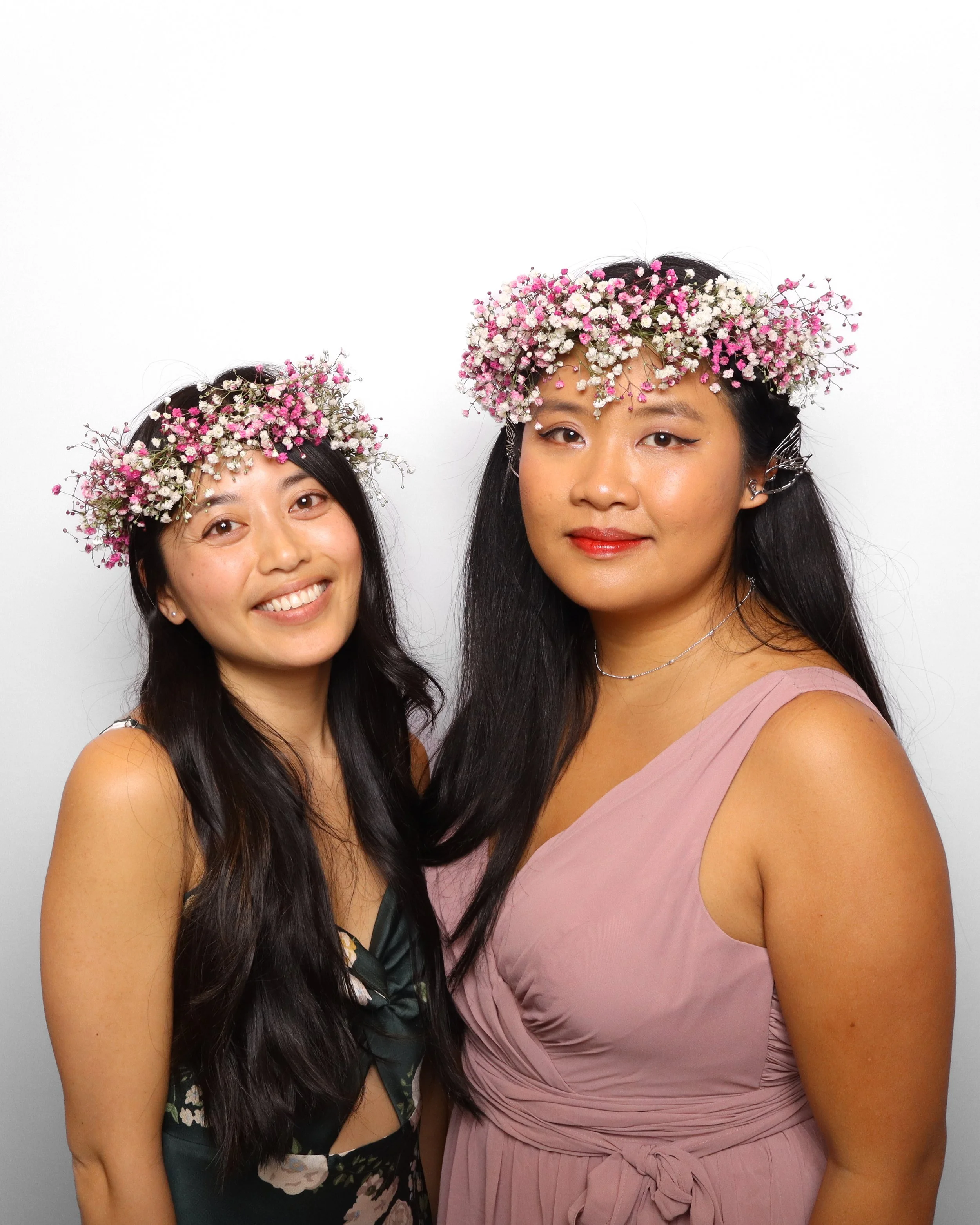 Two women wearing flower crowns made of small pink and white flowers. One woman has dark hair and is wearing a floral dress, the other has long dark hair and is wearing a light pink dress with a bow.