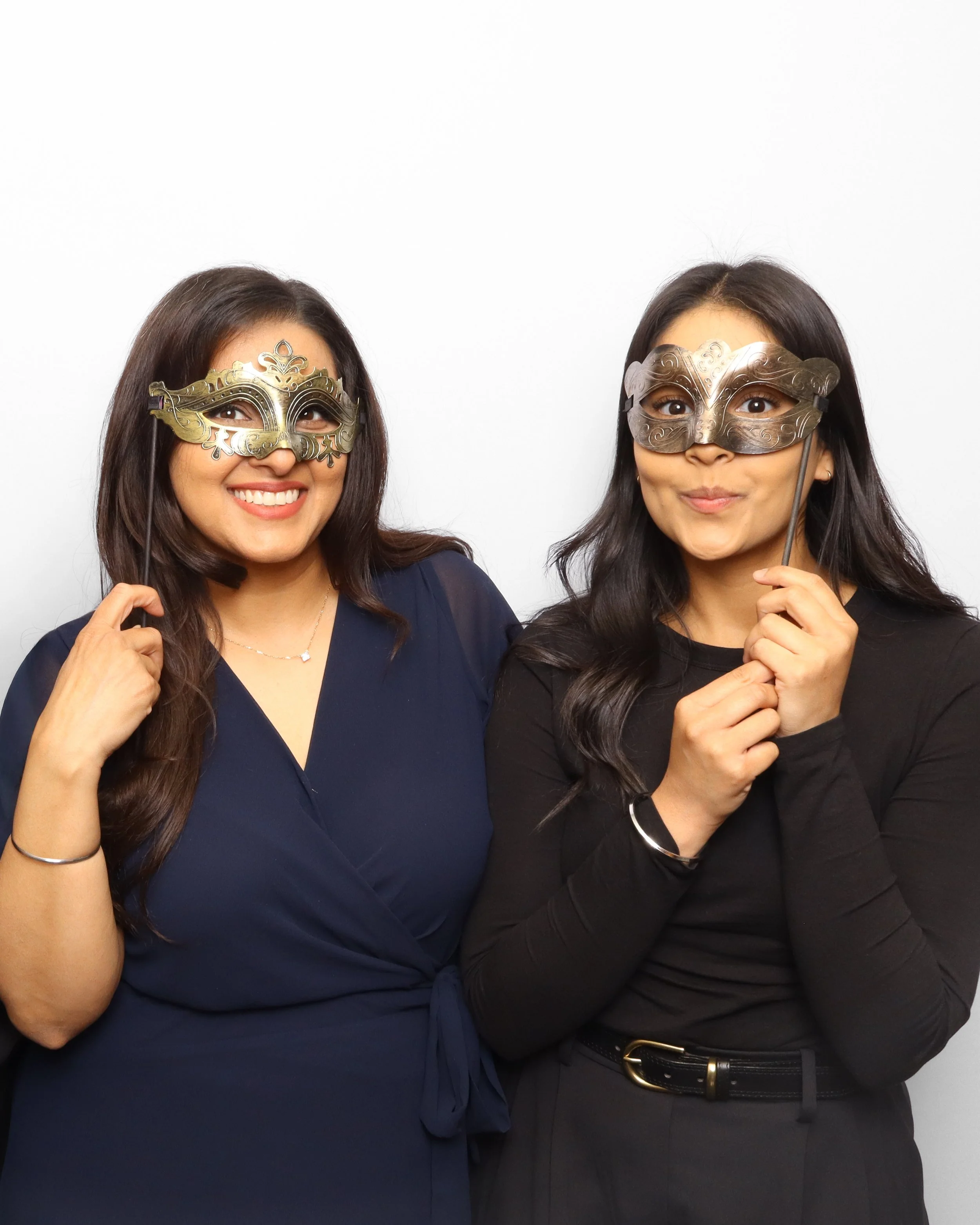 Two women smiling and wearing decorative masks, one in a dark dress and the other in a dark top, against a plain white background.