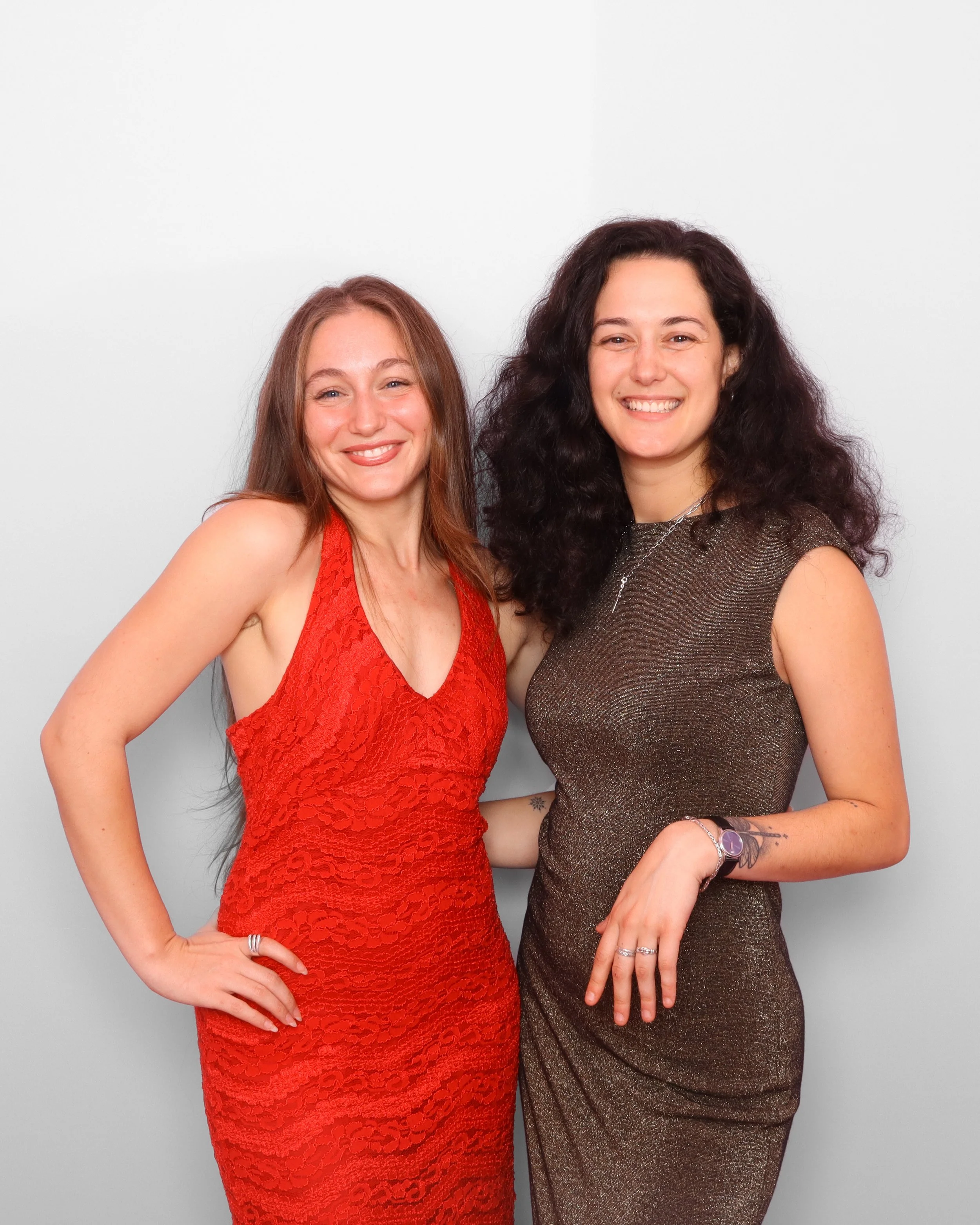 Two women standing close together against a plain white wall, smiling at the camera. The woman on the left has long red hair and is wearing a red lace dress. The woman on the right has long dark curly hair and is wearing a shimmering gold dress.
