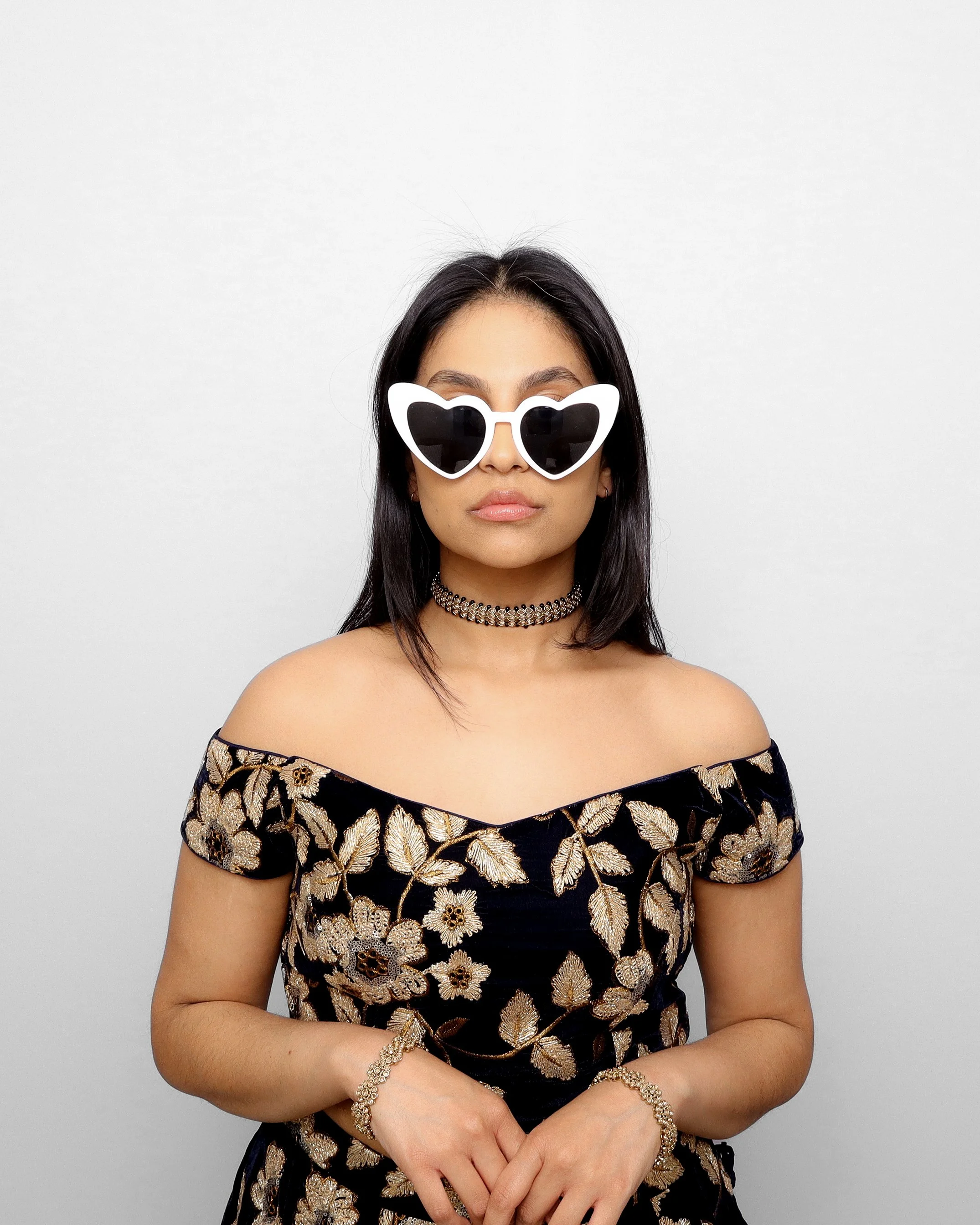 A woman with straight black hair, wearing white heart-shaped sunglasses, a chunky bracelet, and a black off-shoulder dress with gold and beige embroidered floral patterns, stands against a plain gray background.