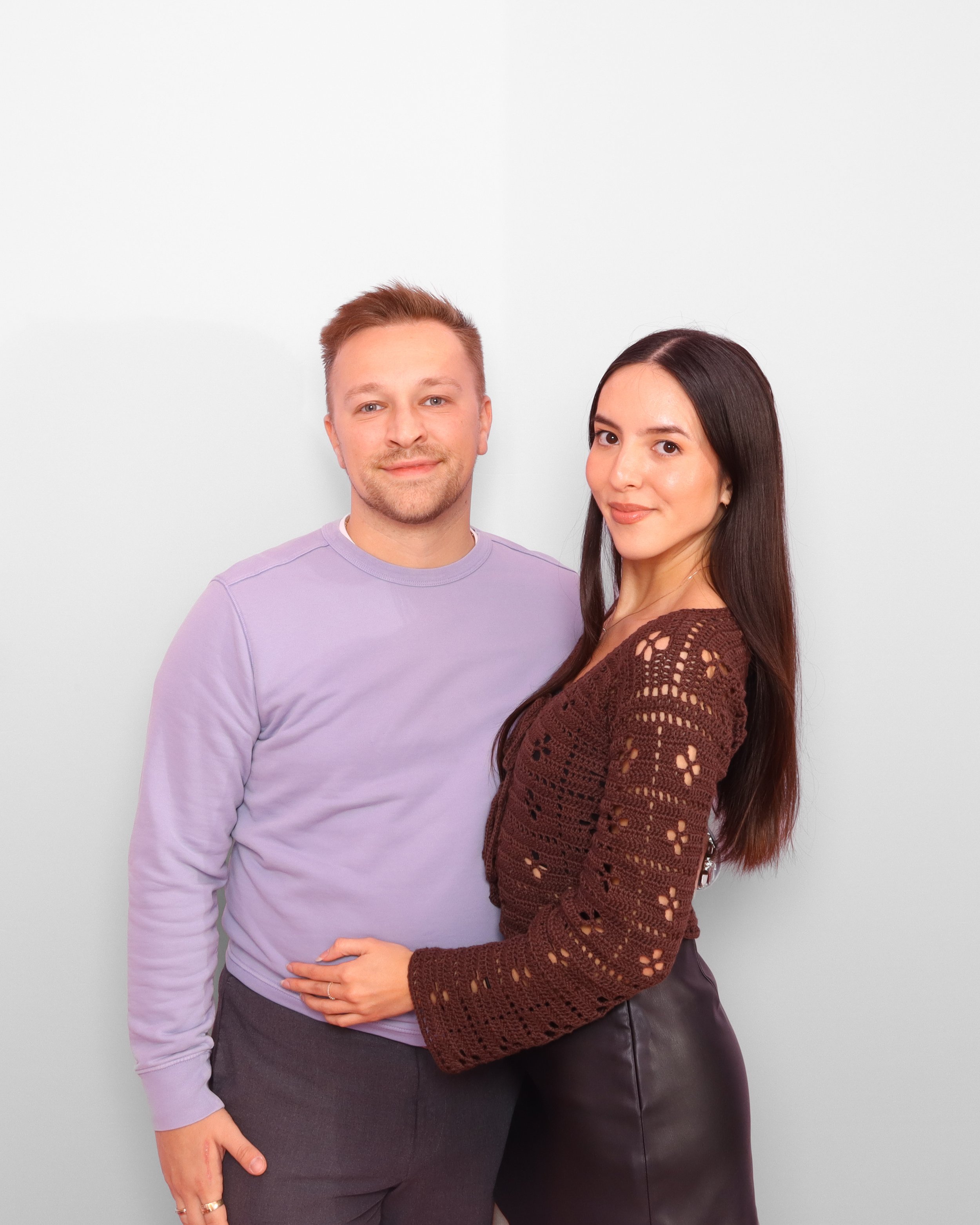 A man and woman stand closely together against a plain white background, with the woman’s arm around the man’s waist. The man has short reddish hair, a slight beard, and is wearing a light purple long sleeve shirt and gray pants. The woman has long d