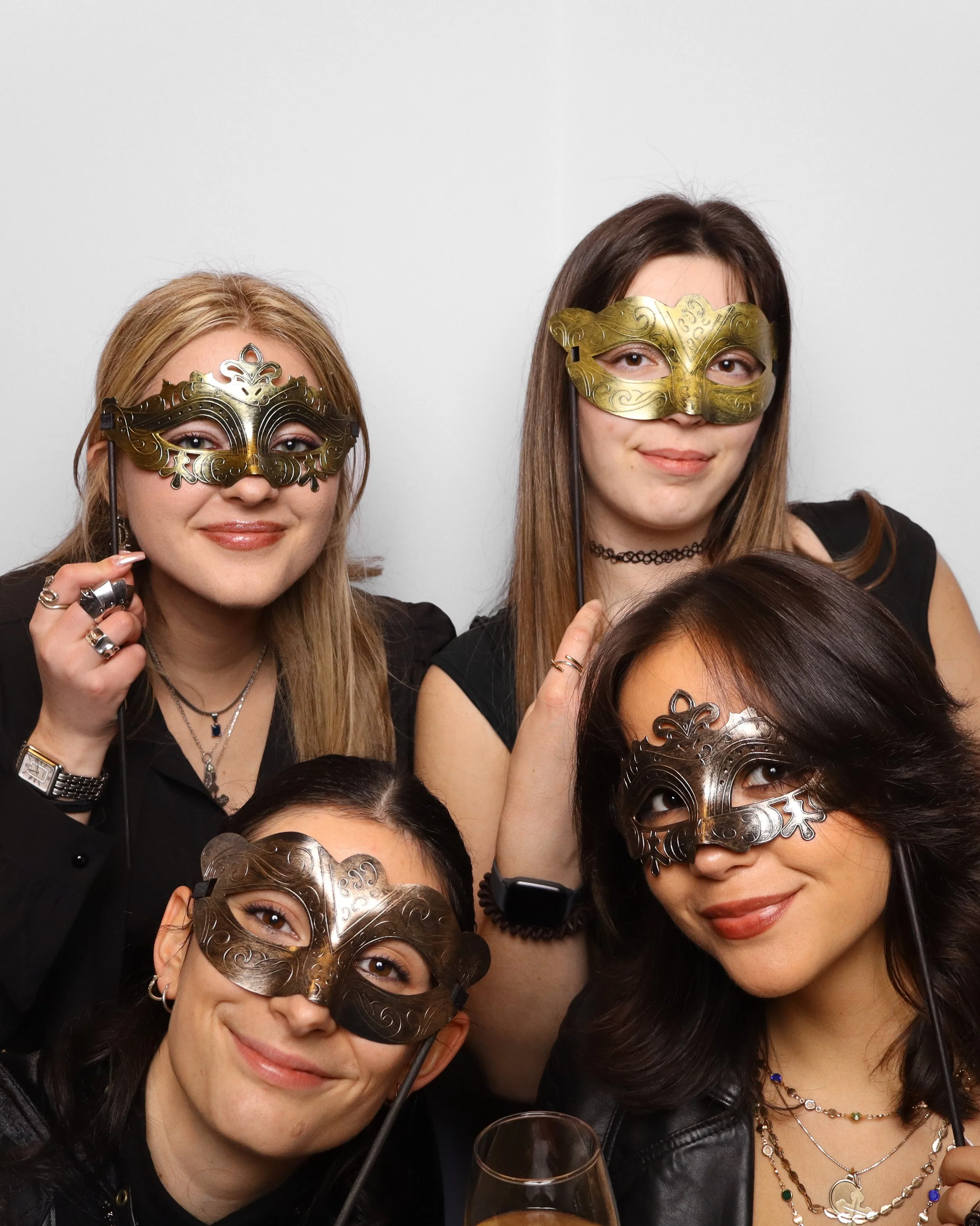 Group of women wearing decorative masks at a celebration or party, smiling and holding glasses.