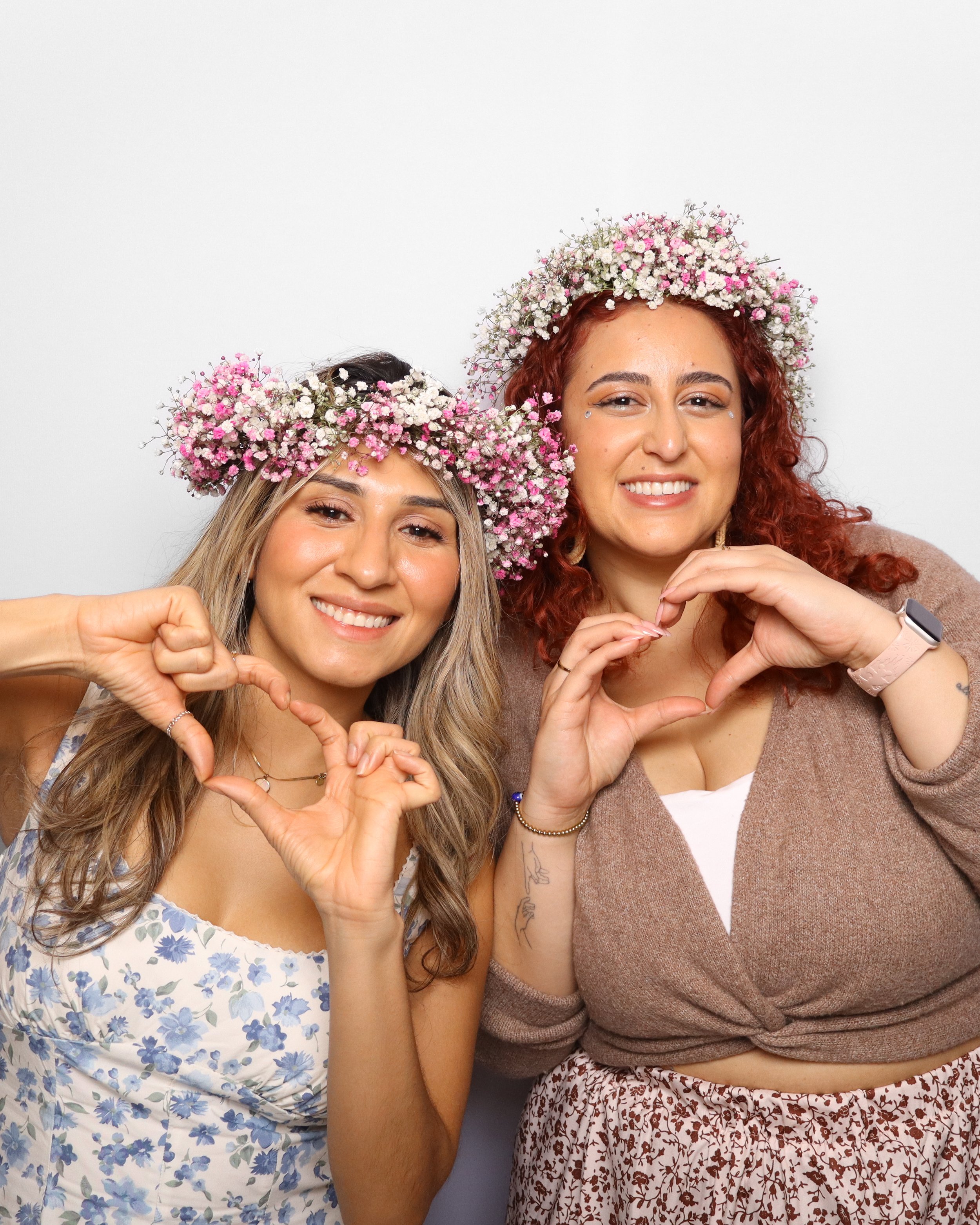 Two women wearing flower crowns made of small pink and white flowers, smiling and forming heart shapes with their hands, standing against a plain white background.