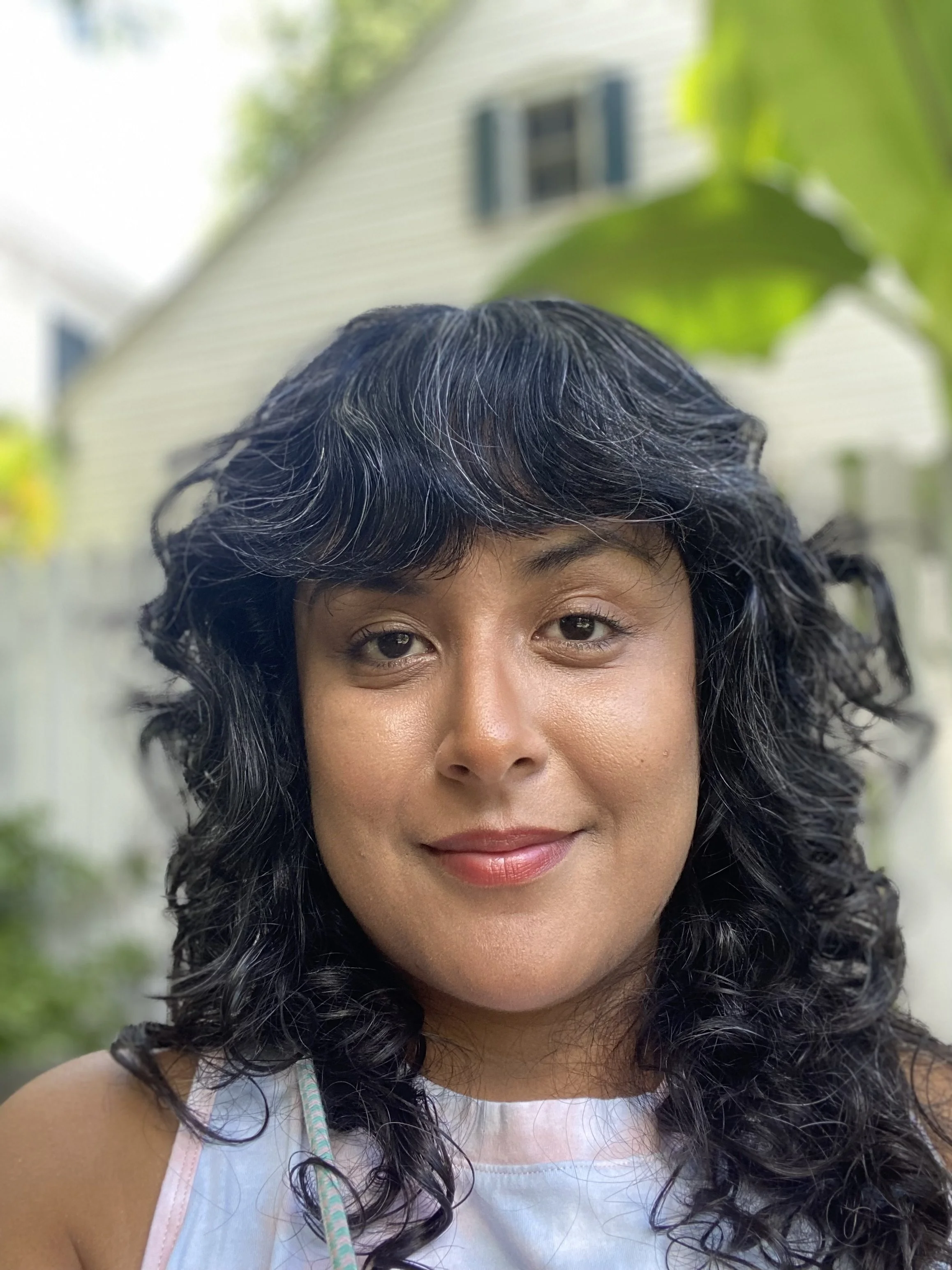 A woman with curly black hair and light makeup smiling outdoors, with a house and green foliage in the background.
