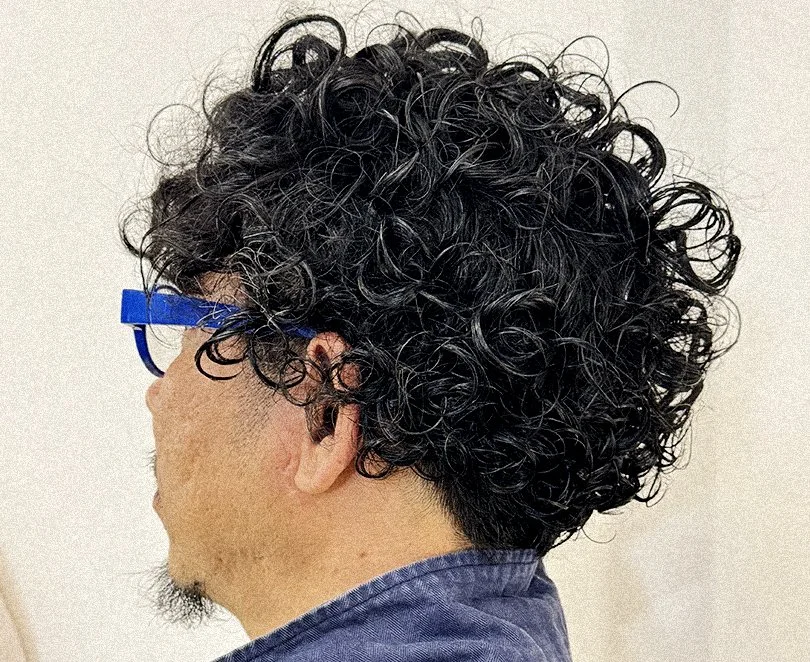 Side view of a person with curly permed hair and blue glasses, wearing a denim shirt.