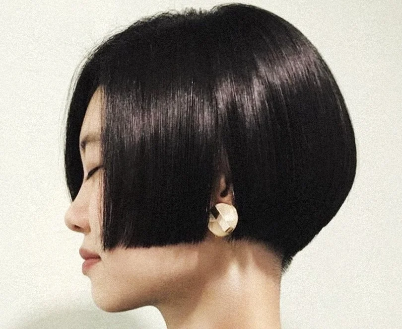 Side profile of woman with a short, asymmetrical bob haircut in dark purple color, wearing large cream-colored earrings.