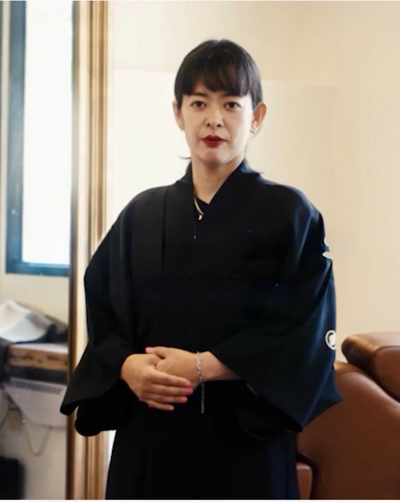 A woman with short dark hair and red lipstick, dressed in a black outfit, standing indoors near a mirror and a window.