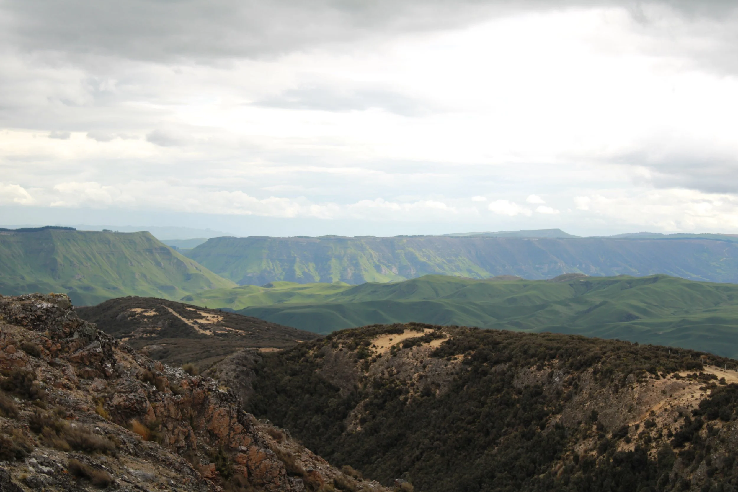 View of a mountain landscape with rugged rocky foreground, rolling green hills, and distant cliffs under a cloudy sky.