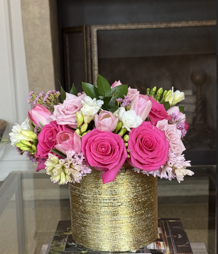 A gold textured flower arrangement vase with pink and white roses, lilies, and other flowers on a glass table in a living room.