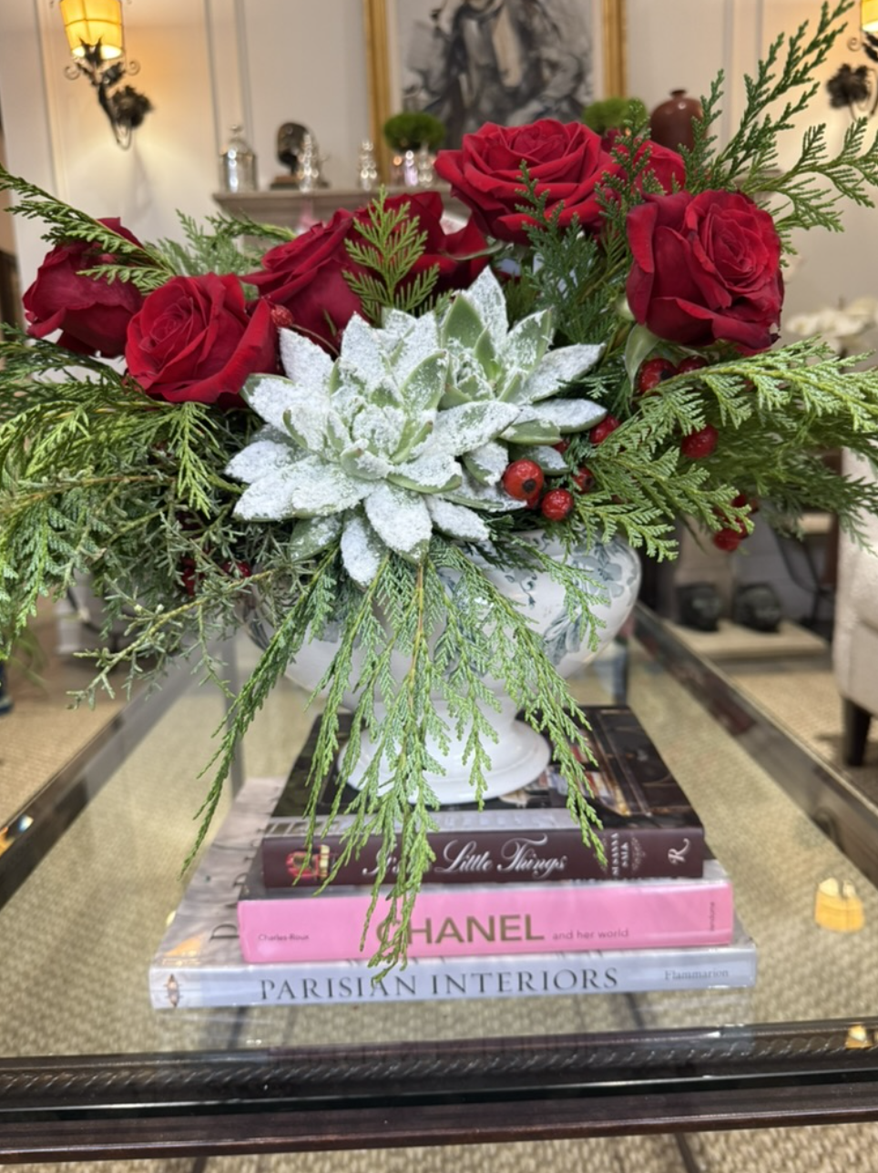 Decorative floral arrangement with red roses, snowy pinecone, and greenery in a white vase, placed on top of fashion and interior design books on a glass table.