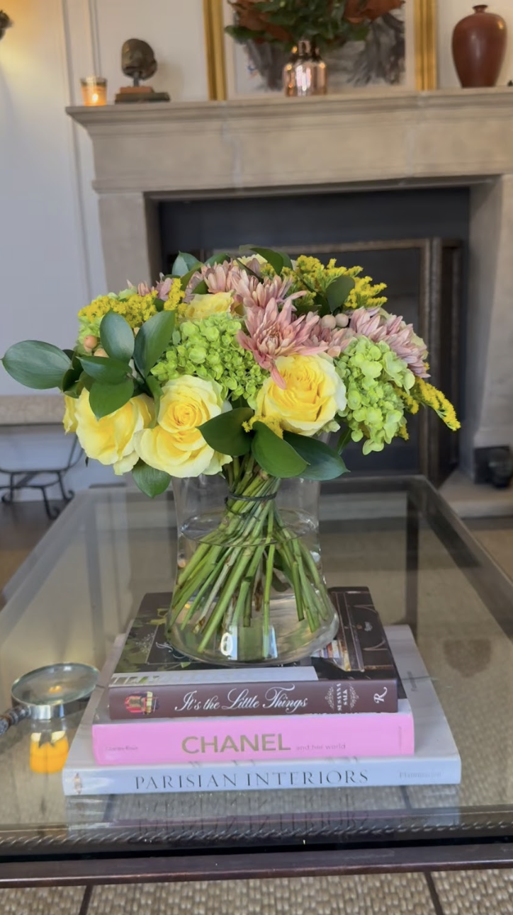 A glass vase filled with yellow roses, pink chrysanthemums, green hydrangeas, and assorted greenery on top of three stacked books on a glass table in front of a fireplace mantel decorated with vases, a sculpture, and candles.